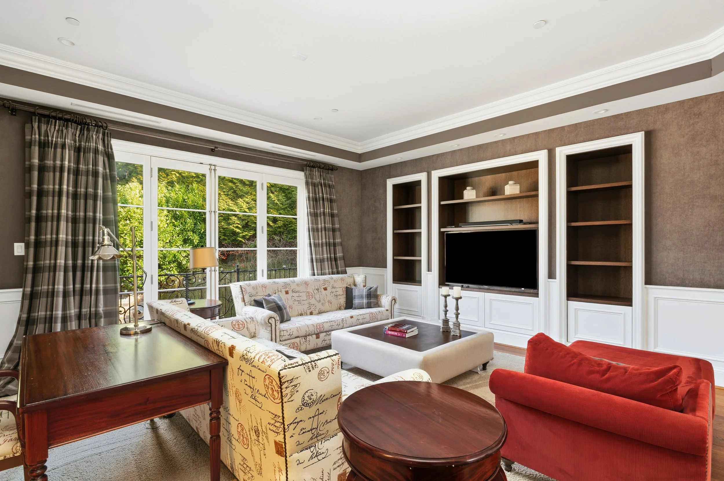Living room with large window, checkered curtains, brown walls, white built-in bookshelves, flat-screen TV, furniture includes red armchair, white loveseat, coffee table, side table with lamp, candle holders, and decorative items.