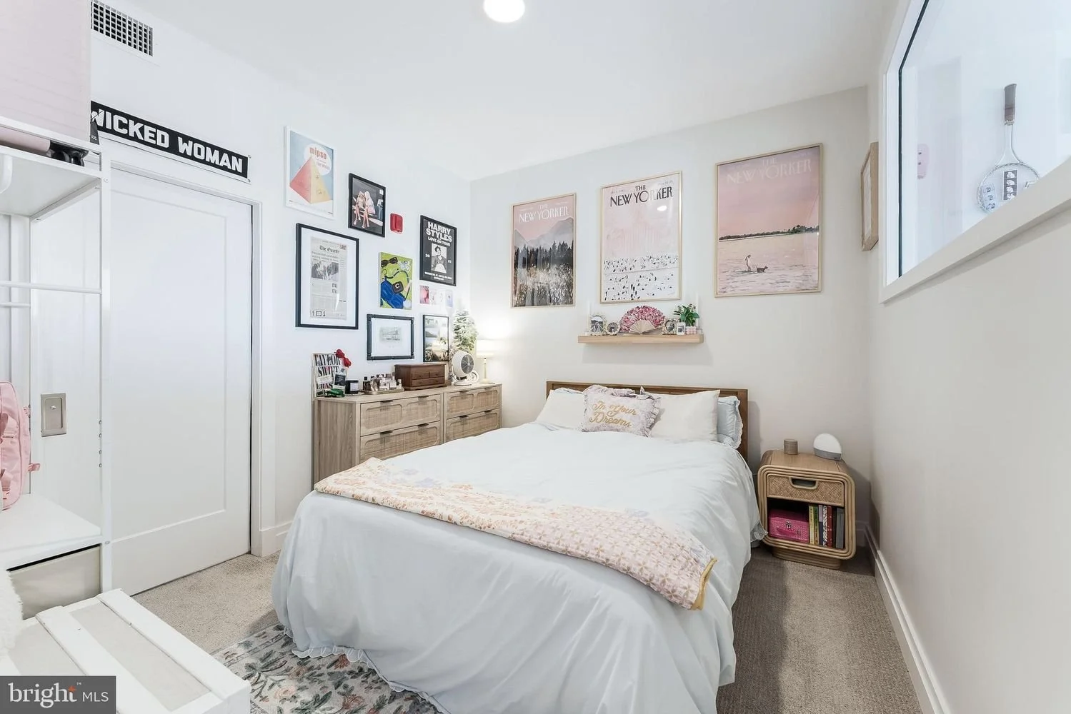 A cozy bedroom with white walls, a white bed with decorative pillows, and a string of artwork above the bed. There is a nightstand with a small lamp and books, a wooden dresser with decorative items, and a wall gallery of framed pictures. A window le