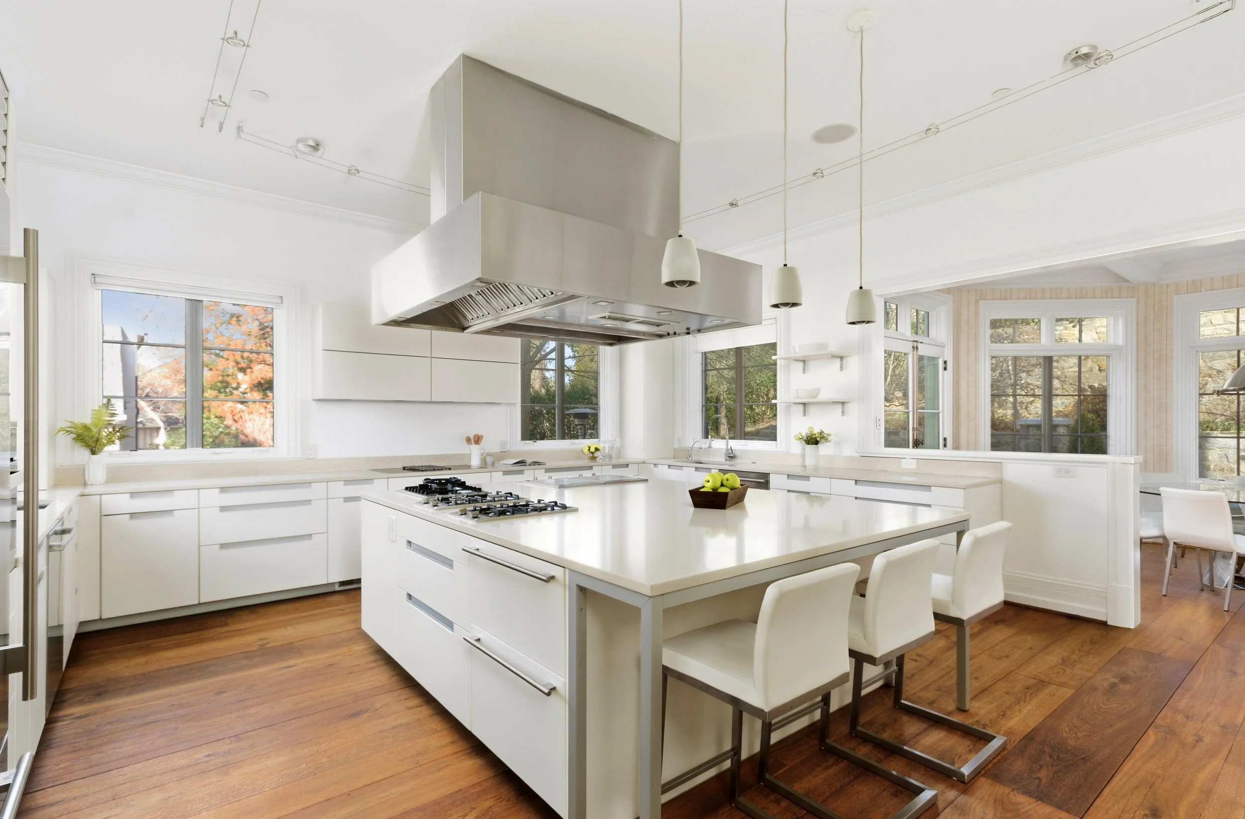 Modern kitchen with white cabinets, a large island with seating, stainless steel appliances, and large windows letting in natural light, with hardwood floors.