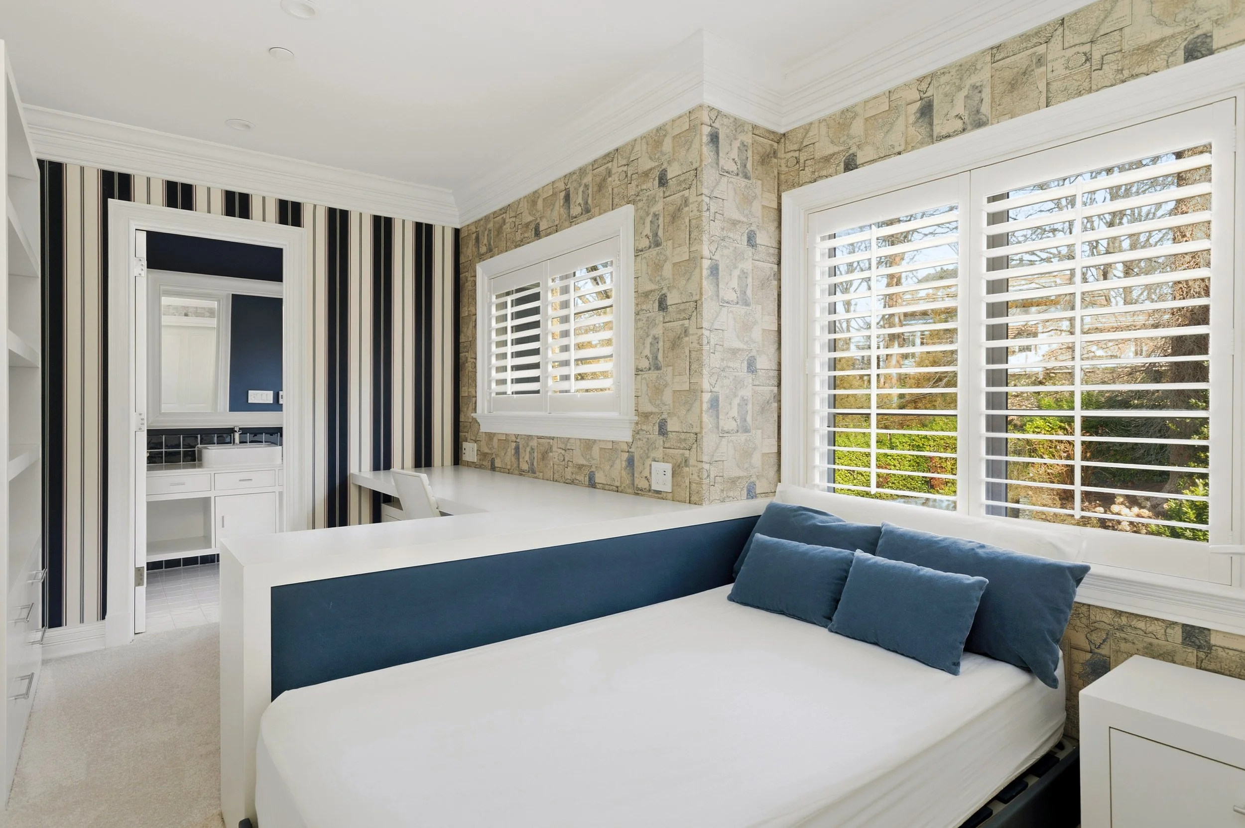 Living room with large windows, white shutters, a blue sofa, and a white bed. The room has a stone accent wall and striped wallpaper. There's a view of trees outside.