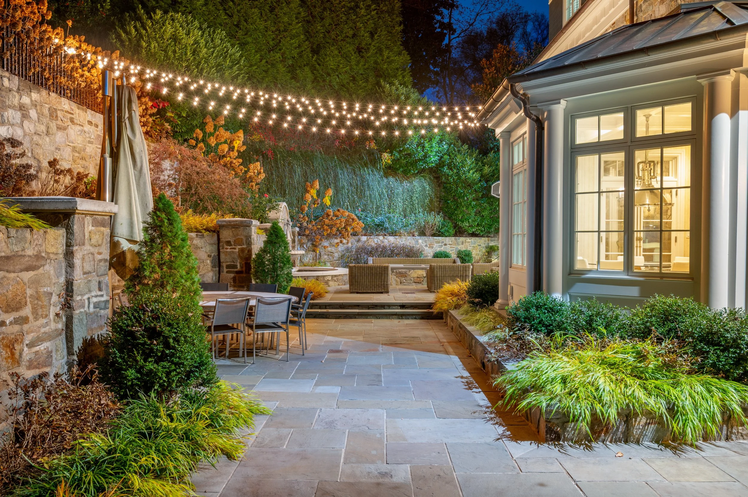 Nighttime backyard with string lights overhead, stone wall on the left, a patio area with chairs, and lush green plants and trees surrounding it, with a house with large windows illuminated on the right.
