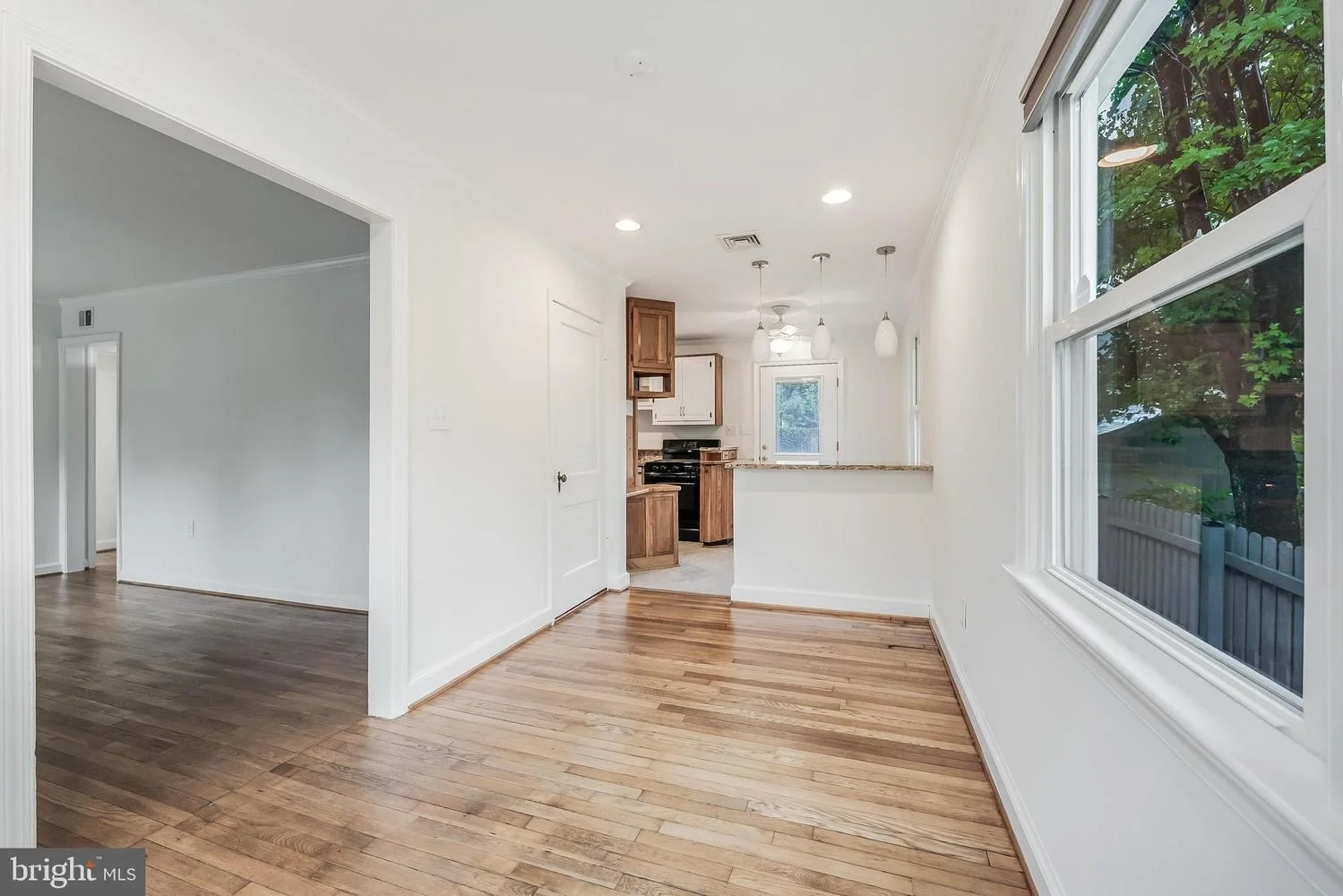 Empty dining area with hardwood floors, white walls, large window, view of a backyard with trees, open to a kitchen with wooden cabinets, granite countertop, black stove, and pendant lighting.
