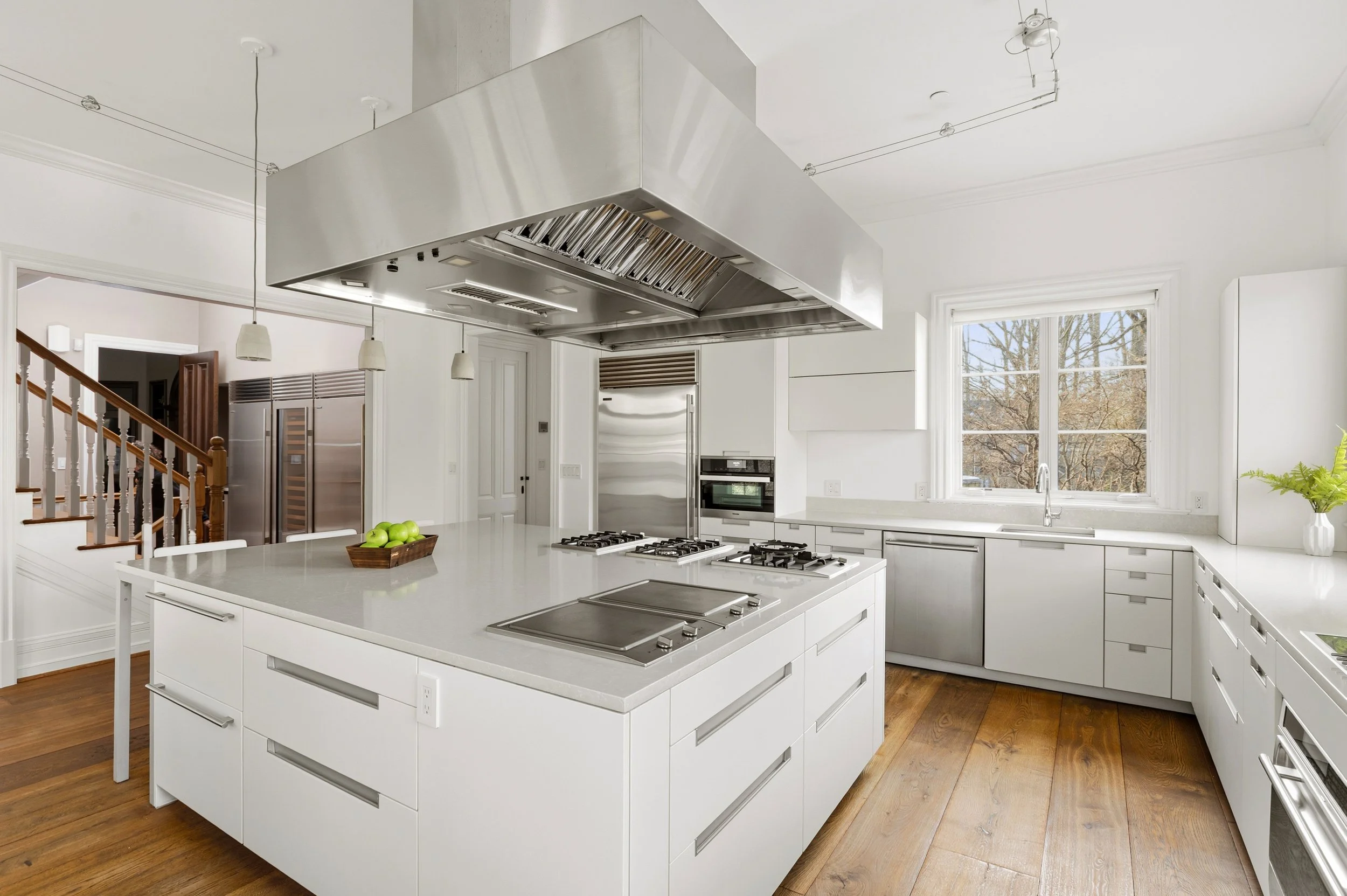 Modern white kitchen with island, built-in stainless steel appliances, large window, and a stainless steel range hood above the island.