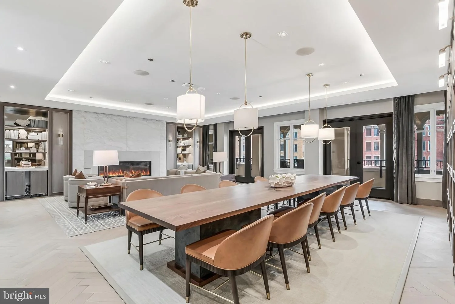 Modern living room with a large wooden dining table, tan chairs, a gray sofa, a fireplace, built-in bookshelves, windows with curtains, and ceiling pendant lights.