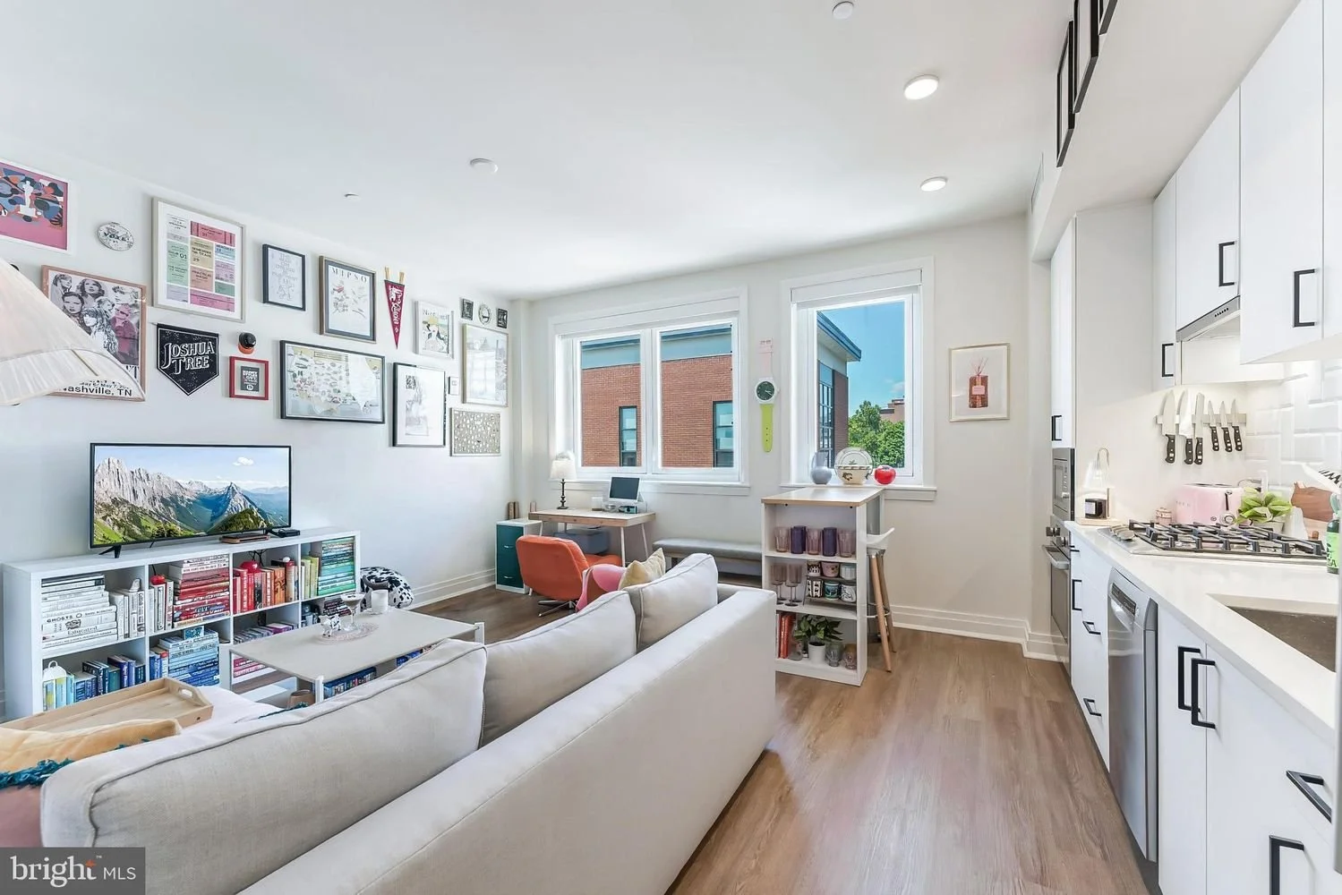 Living room with a white sofa, wall art, a TV, a desk with a computer, and a second window with a view of neighboring buildings. Open kitchen with white cabinets and black handles, a gas stove, and wooden flooring.