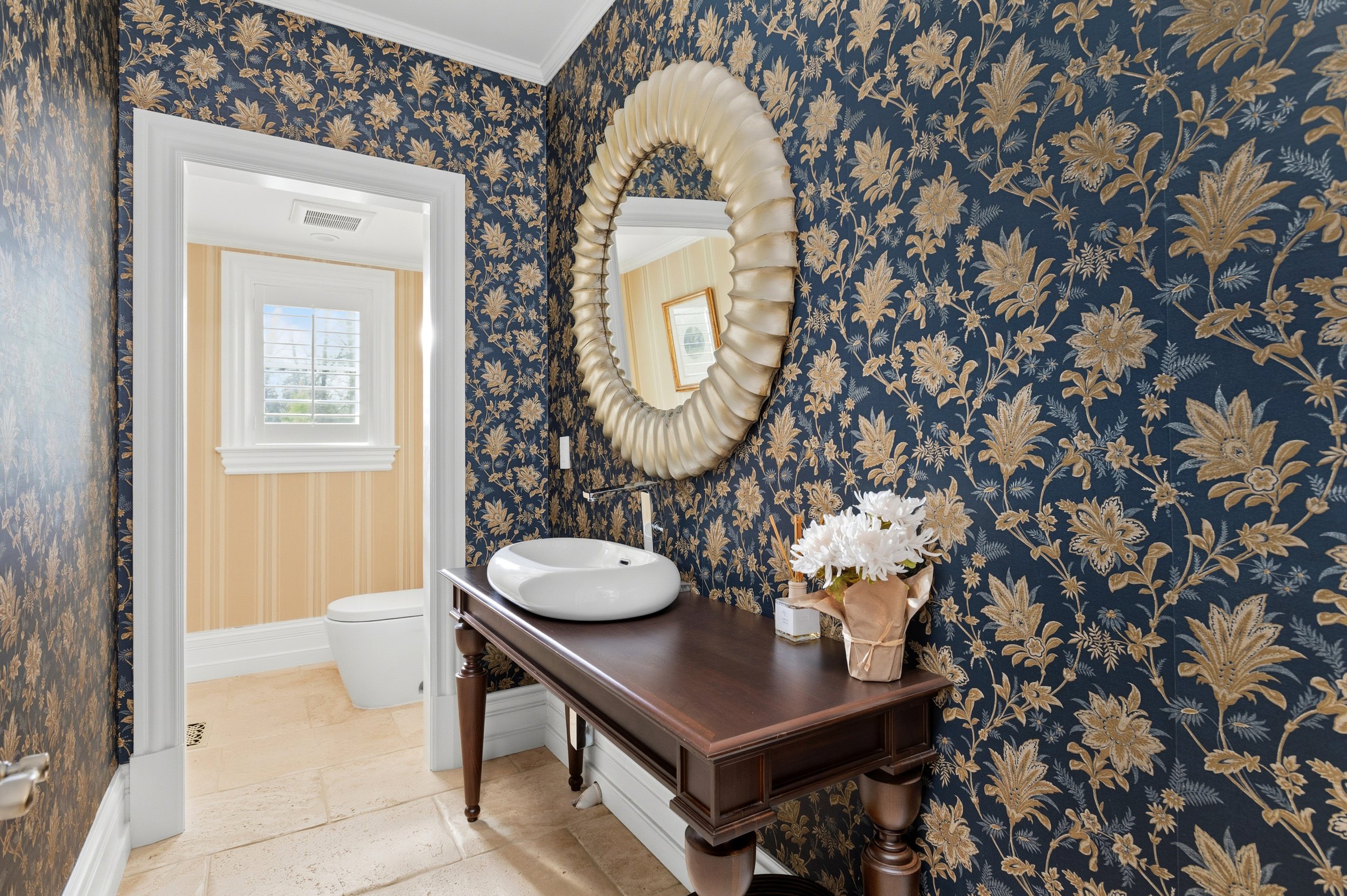A bathroom with blue floral wallpaper, a large gold-framed mirror, a dark wood vanity with a round vessel sink, a white toilet, a window with shutters, and a vase of white flowers on the vanity.