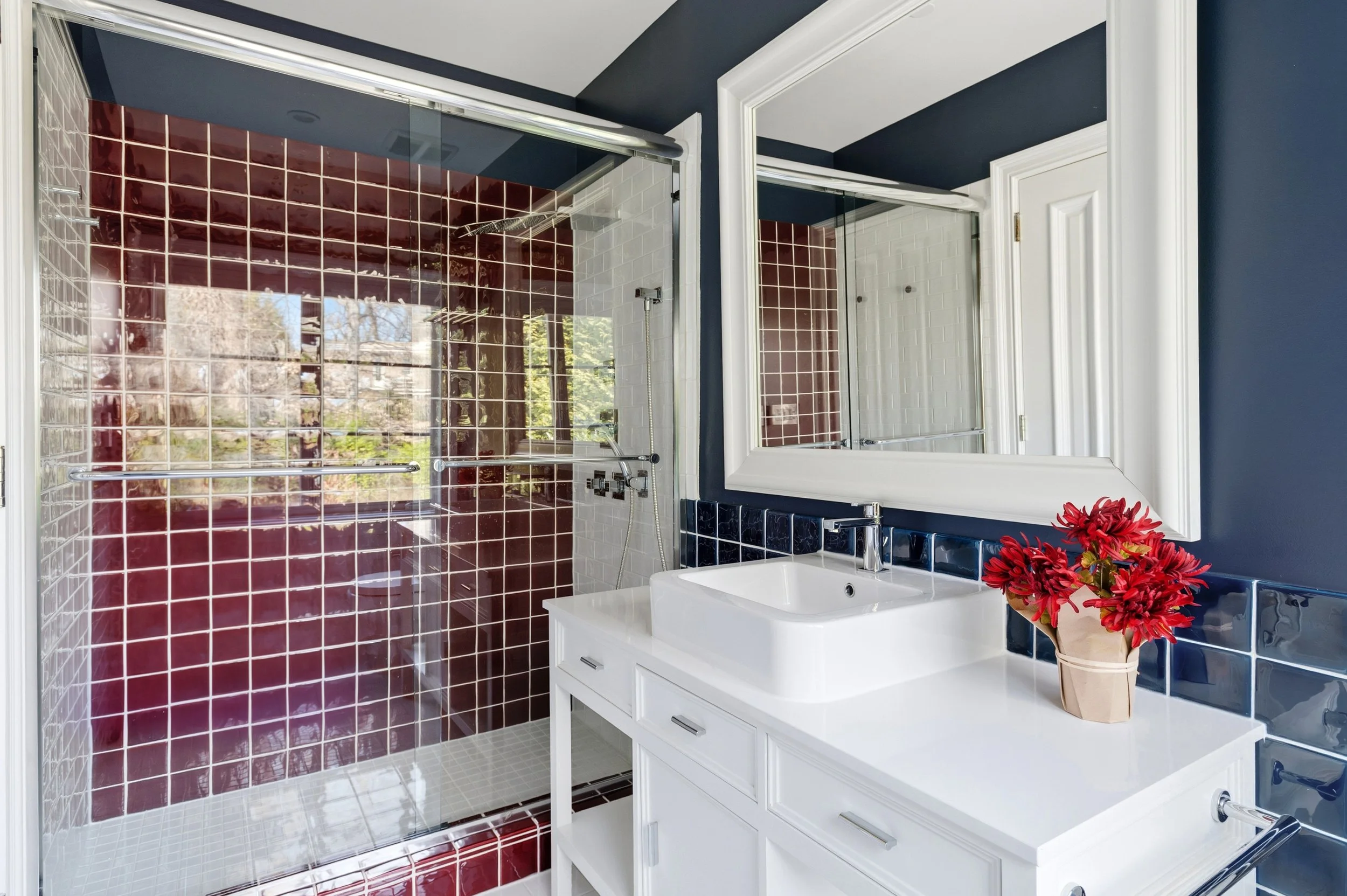 Bathroom with navy blue walls, white vanity with drawers, white sink, large mirror, and a bouquet of red flowers in a paper bag on the vanity. The shower area has red and white tiled walls and a glass door.
