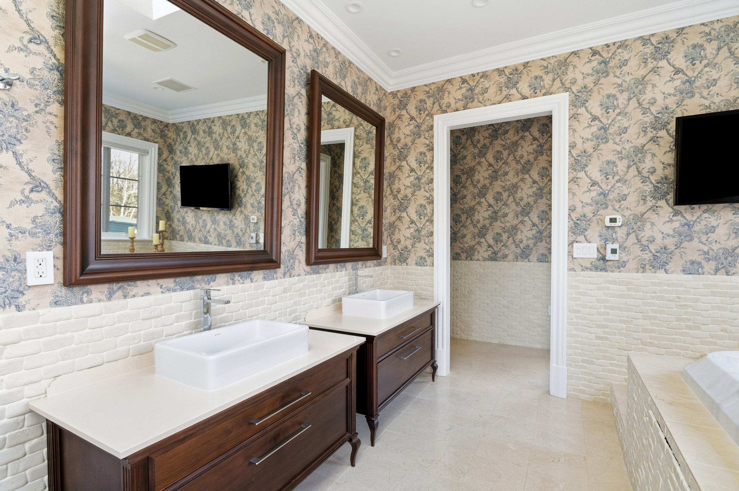 Bathroom with two sinks, wooden vanity, large wall mirrors, floral wallpaper, window, flat-screen TV, thermostats, and beige tiled walls.