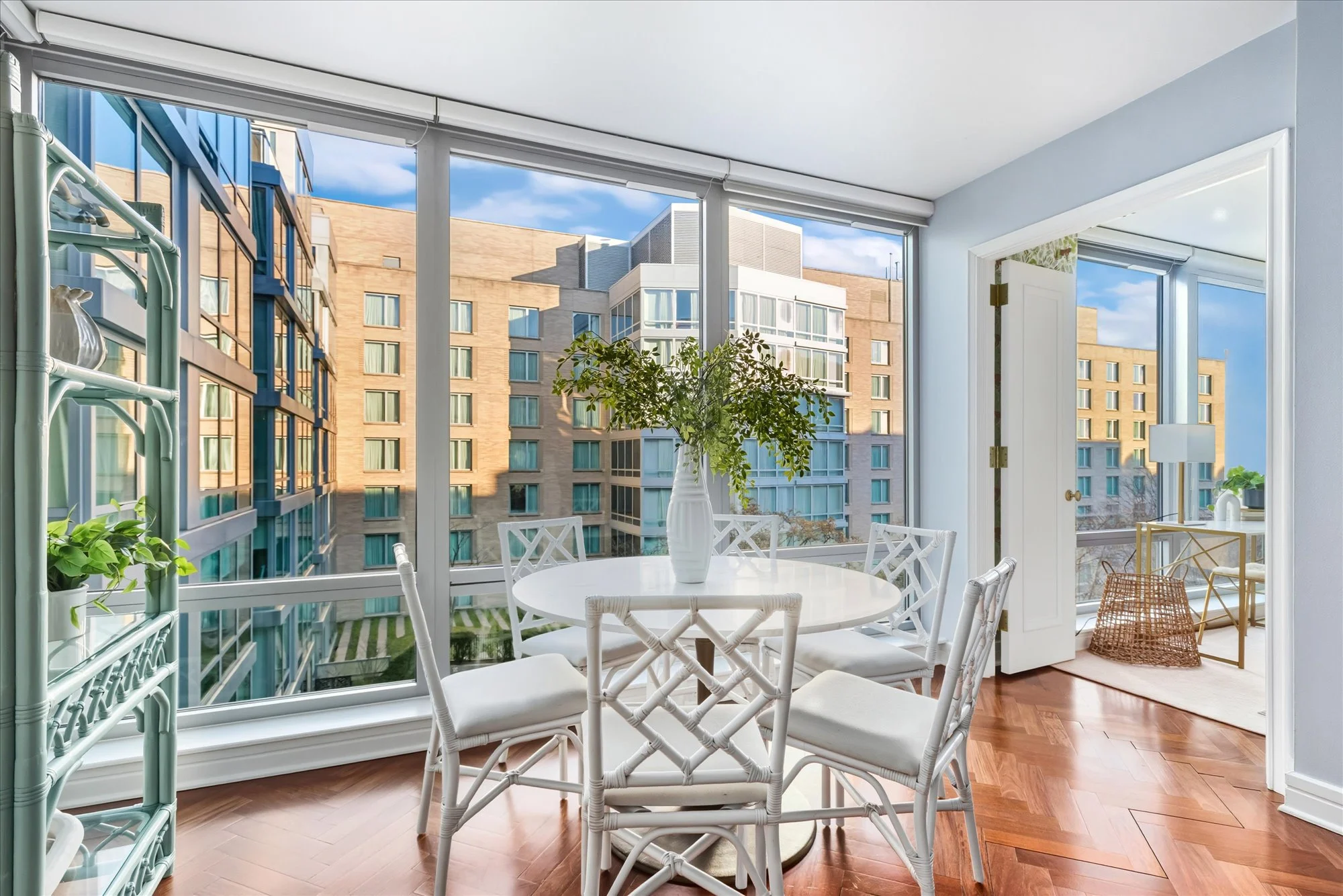 Sunlit enclosed balcony with white wicker chairs and a round table with a white vase and green plant. Large windows reveal surrounding modern apartment buildings and a blue sky.