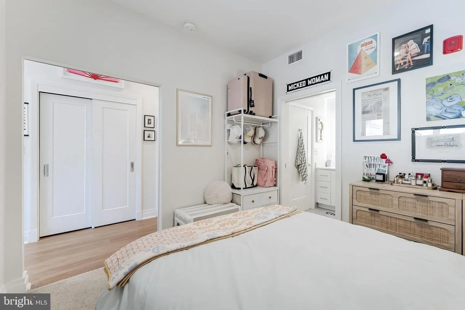 A cozy bedroom with a white bed, wall art, and storage. Open door reveals a bathroom. Decorations include framed pictures, a sign that reads 'Wicked Woman', and a black and white art frame. A dresser with decorative items is on the right.