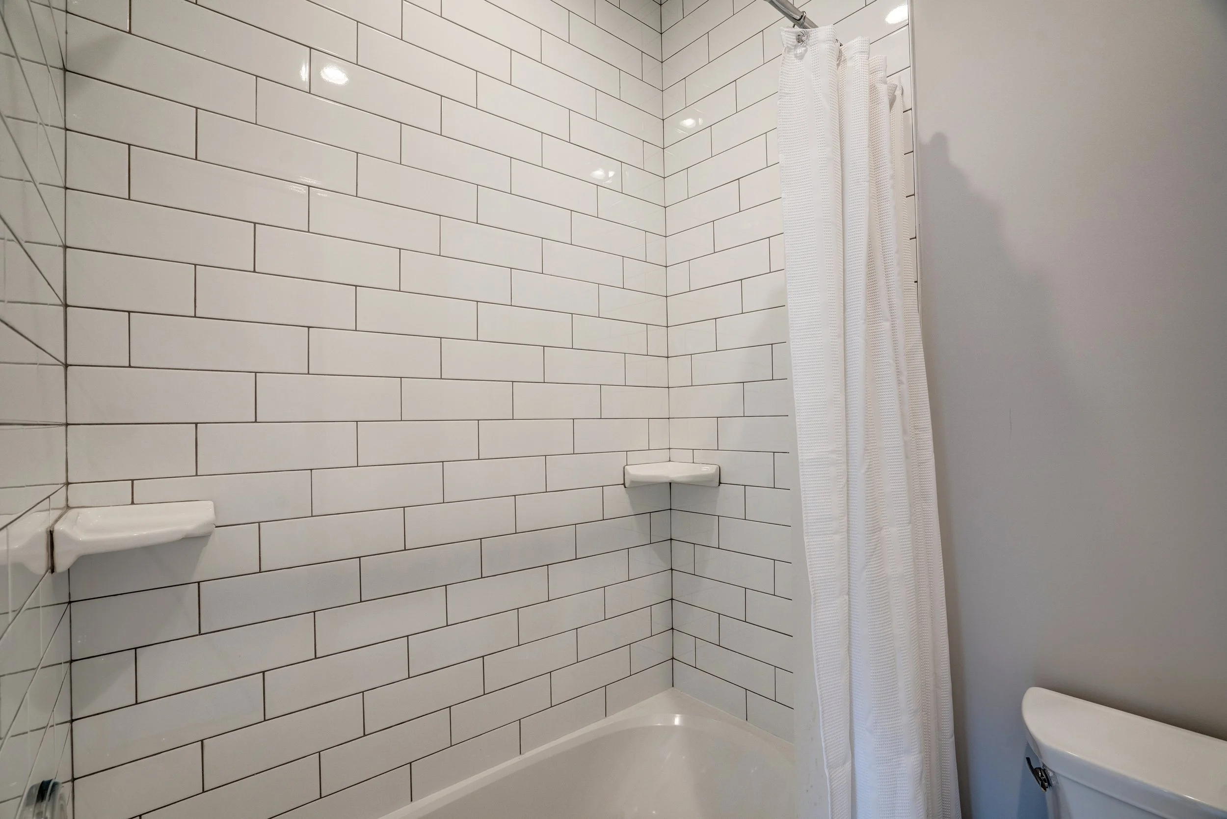 A bathroom with white subway tile walls, a white shower curtain, and a toilet beside the shower.