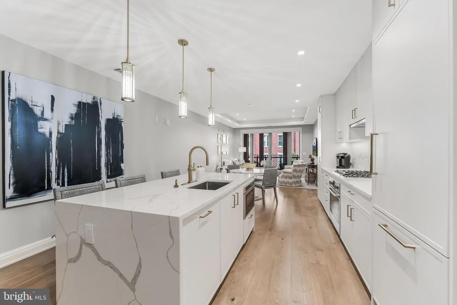 Modern open-concept kitchen with white cabinetry, marble island, and hanging pendant lights, overlooking a living area with art and seating.