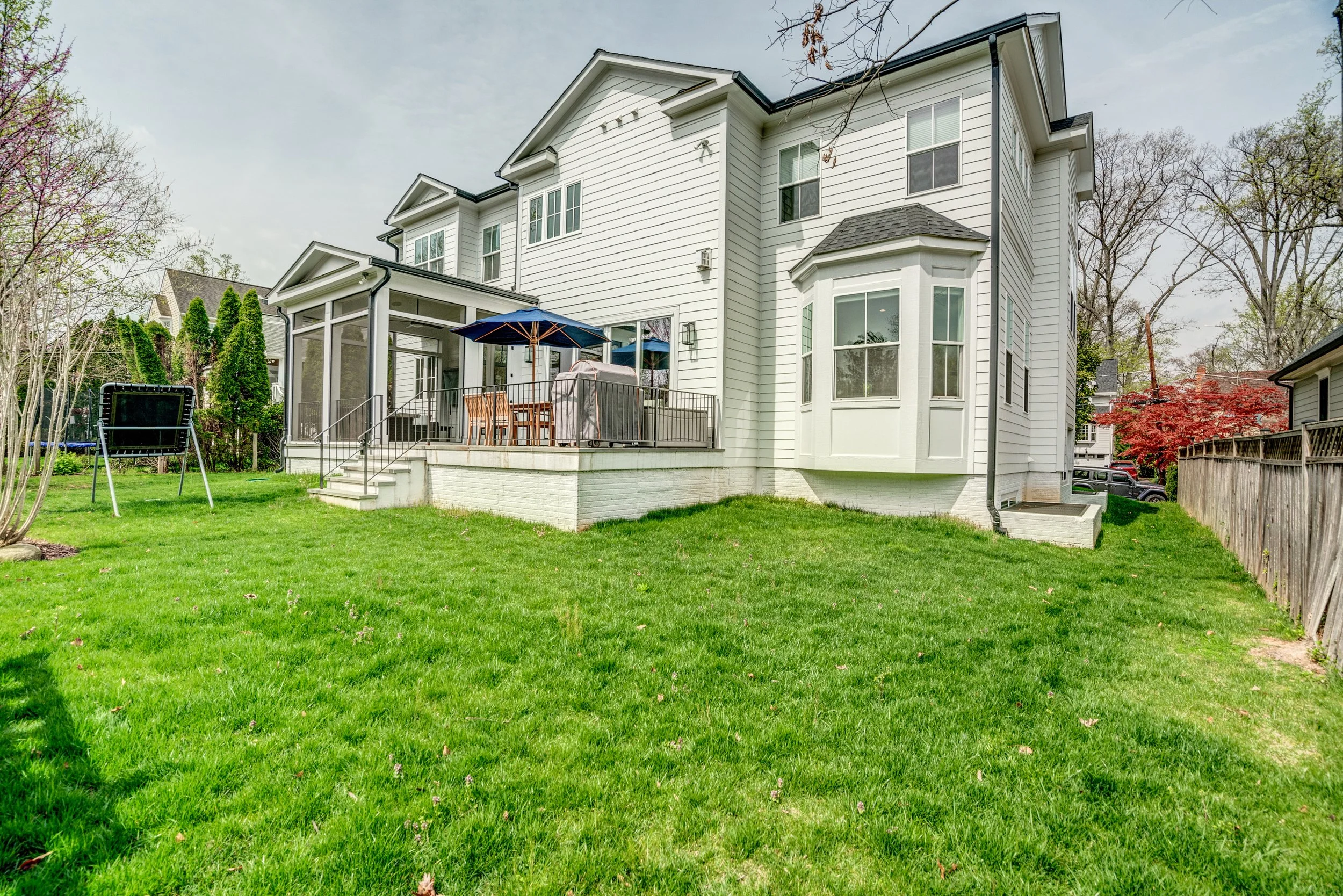 Backyard of a white, multi-story house with large windows, a patio with outdoor furniture, umbrellas, and a trampoline, surrounded by green lawn, trees, and neighboring houses.