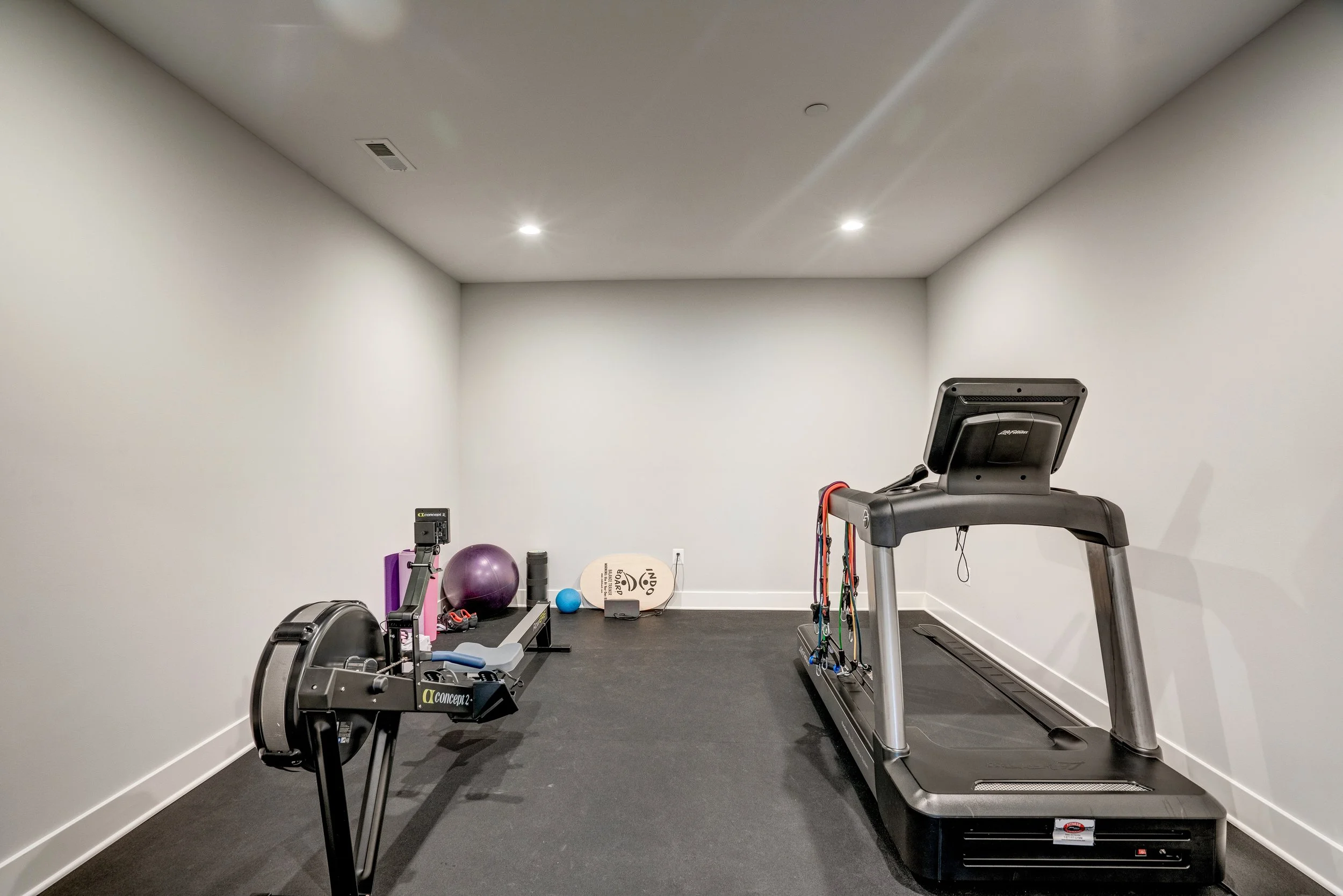 A small home gym with a treadmill, rowing machine, exercise balls, and various workout equipment against bare white walls and black flooring.