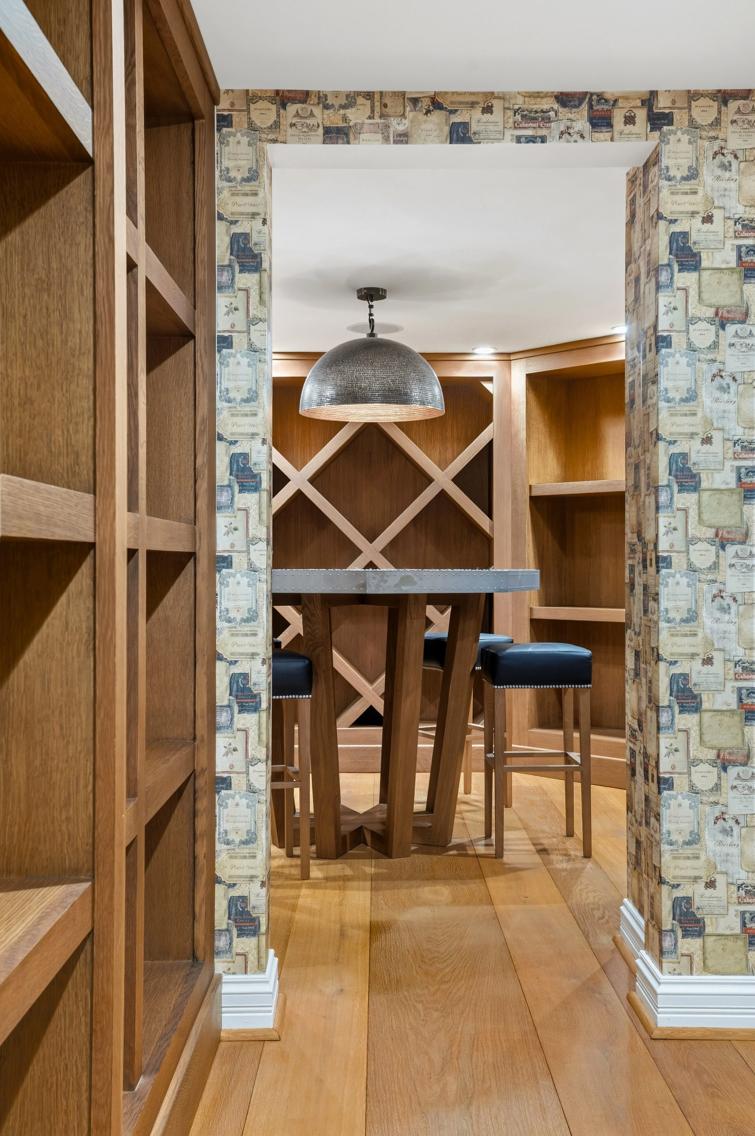 Wooden dining table with black cushioned barstools in a cozy corner, framed by wallpapered walls with vintage card prints, wooden shelving, and a metallic pendant light overhead.