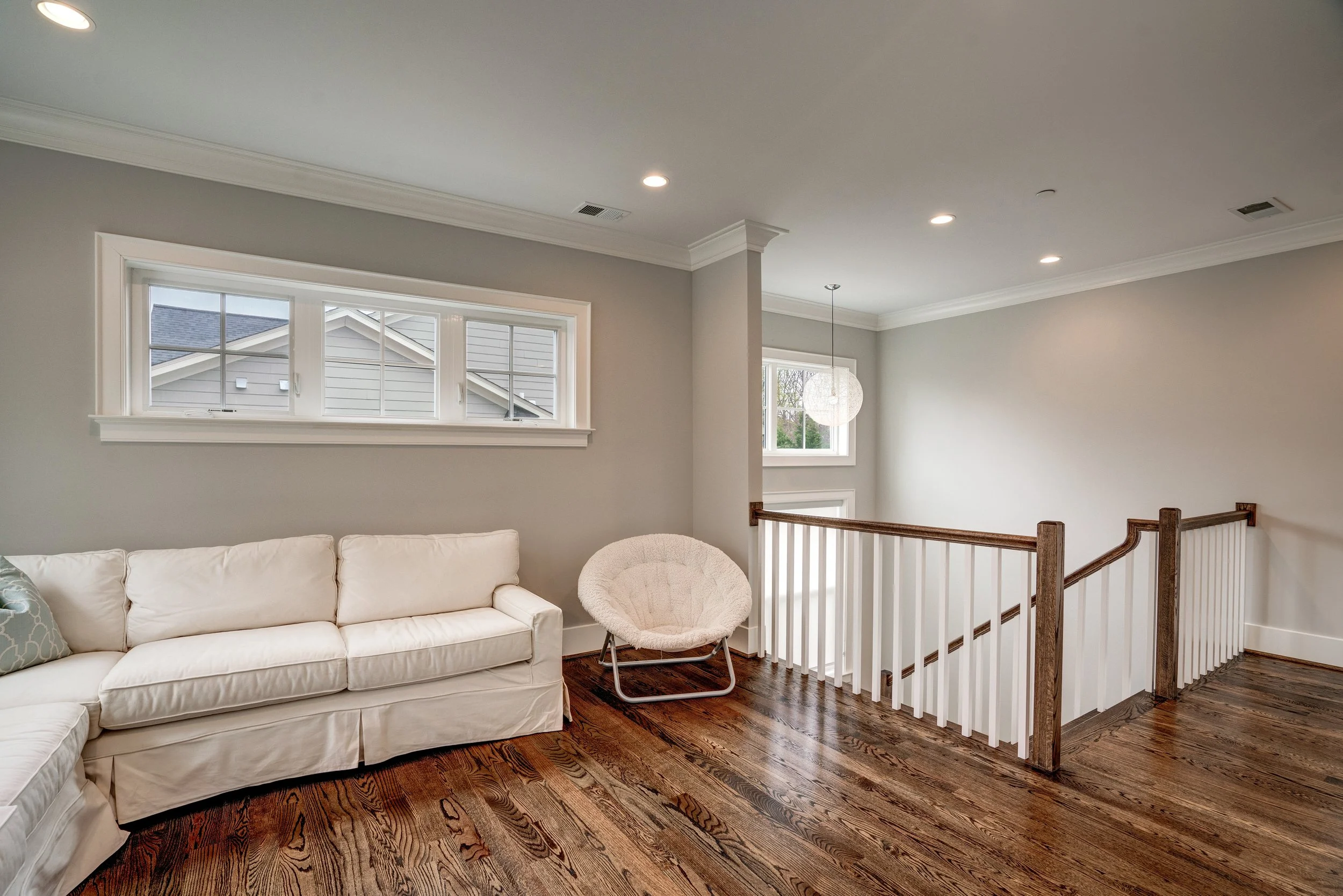 Living room with white sofa, a cozy armchair, hardwood floors, large windows, a staircase with wooden handrails, and a pendant light