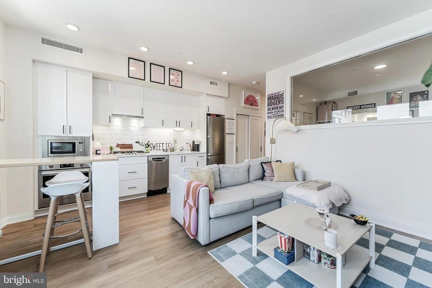 Open living room and kitchen area with white cabinetry, stainless steel appliances, a light gray sofa with pillows, a white coffee table, and decorative artwork on the walls.