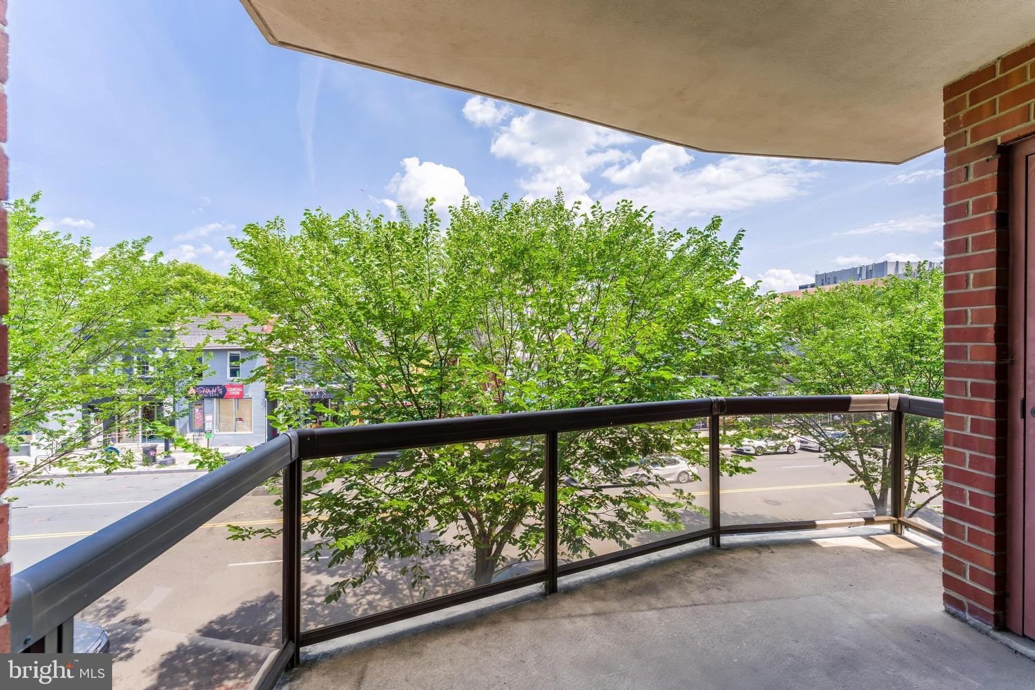 View from a balcony overlooking a street lined with trees and shops, with blue sky and clouds above.