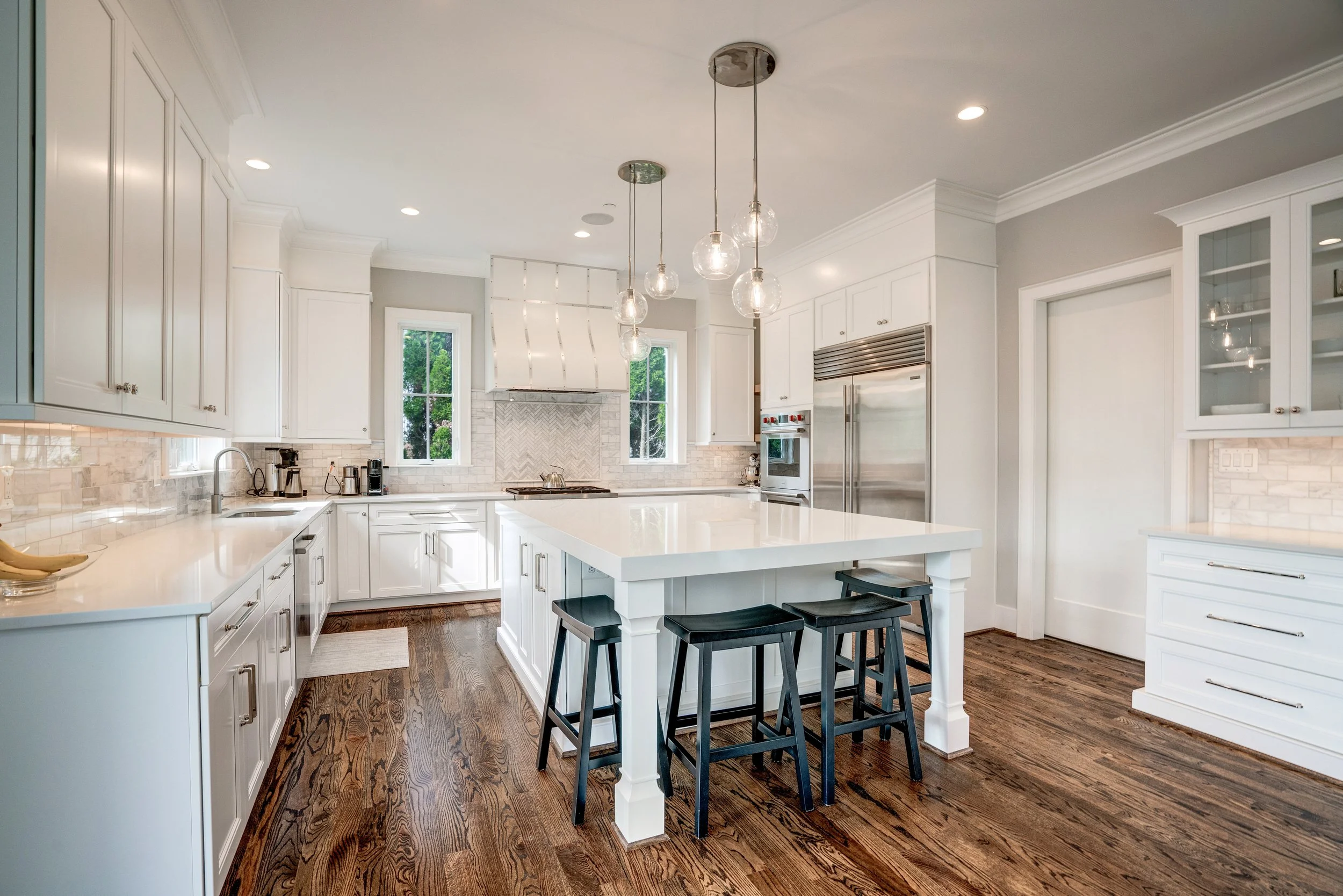 Bright kitchen with white cabinets, a central island with seating, and pendant lights hanging from the ceiling. Windows show greenery outside, and hardwood floors are visible.