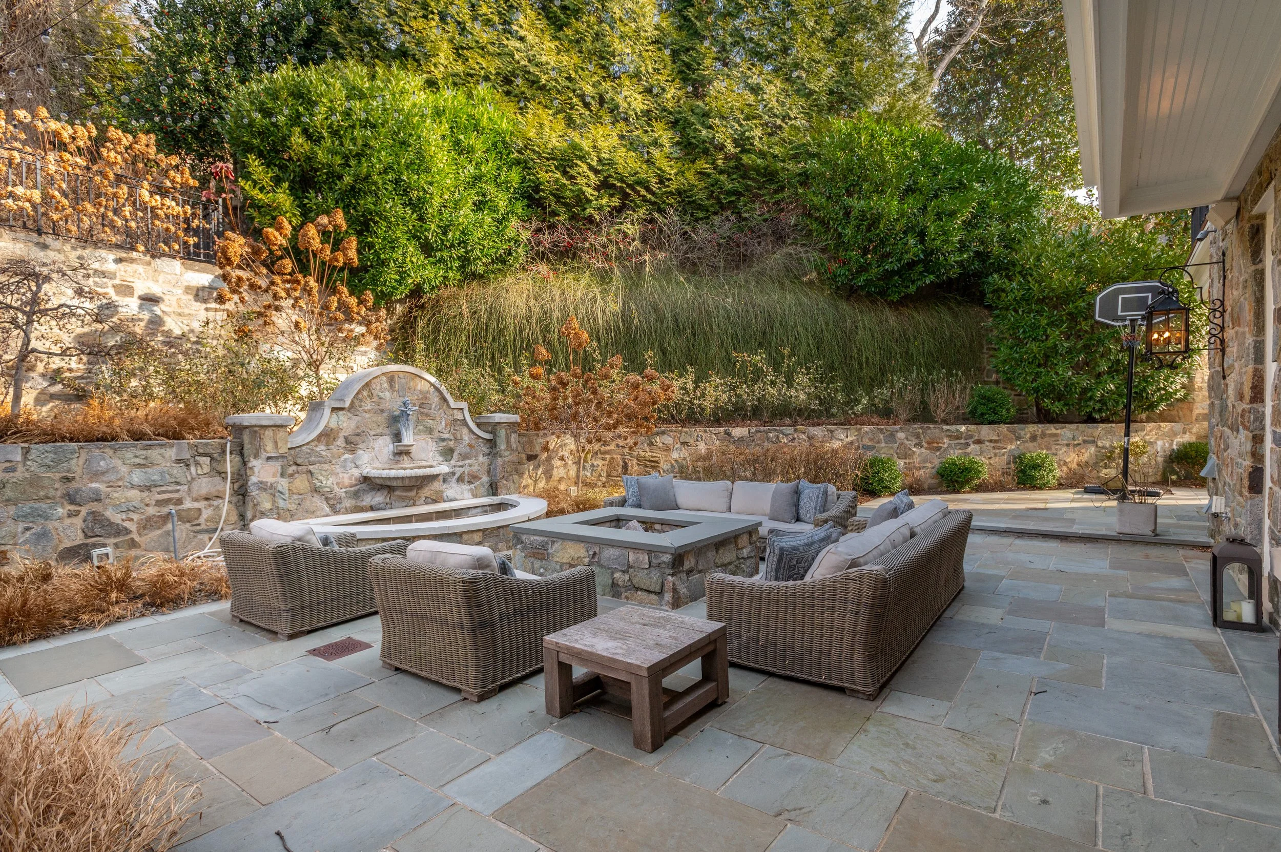 A backyard patio area with outdoor furniture including a sofa, chairs, and coffee tables made of wicker and wood. There is a stone wall with a fountain and lush green trees and shrubs in the background. A basketball hoop and a street lamp are attache