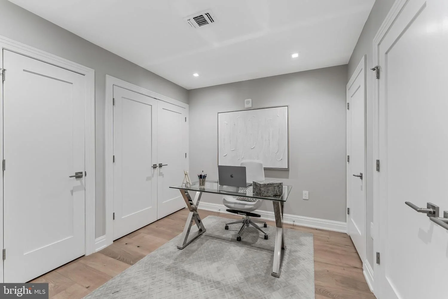 Modern home office with a glass desk, white ergonomic chair, laptop, and decor, with light gray walls, hardwood floors, white doors, and recessed lighting.