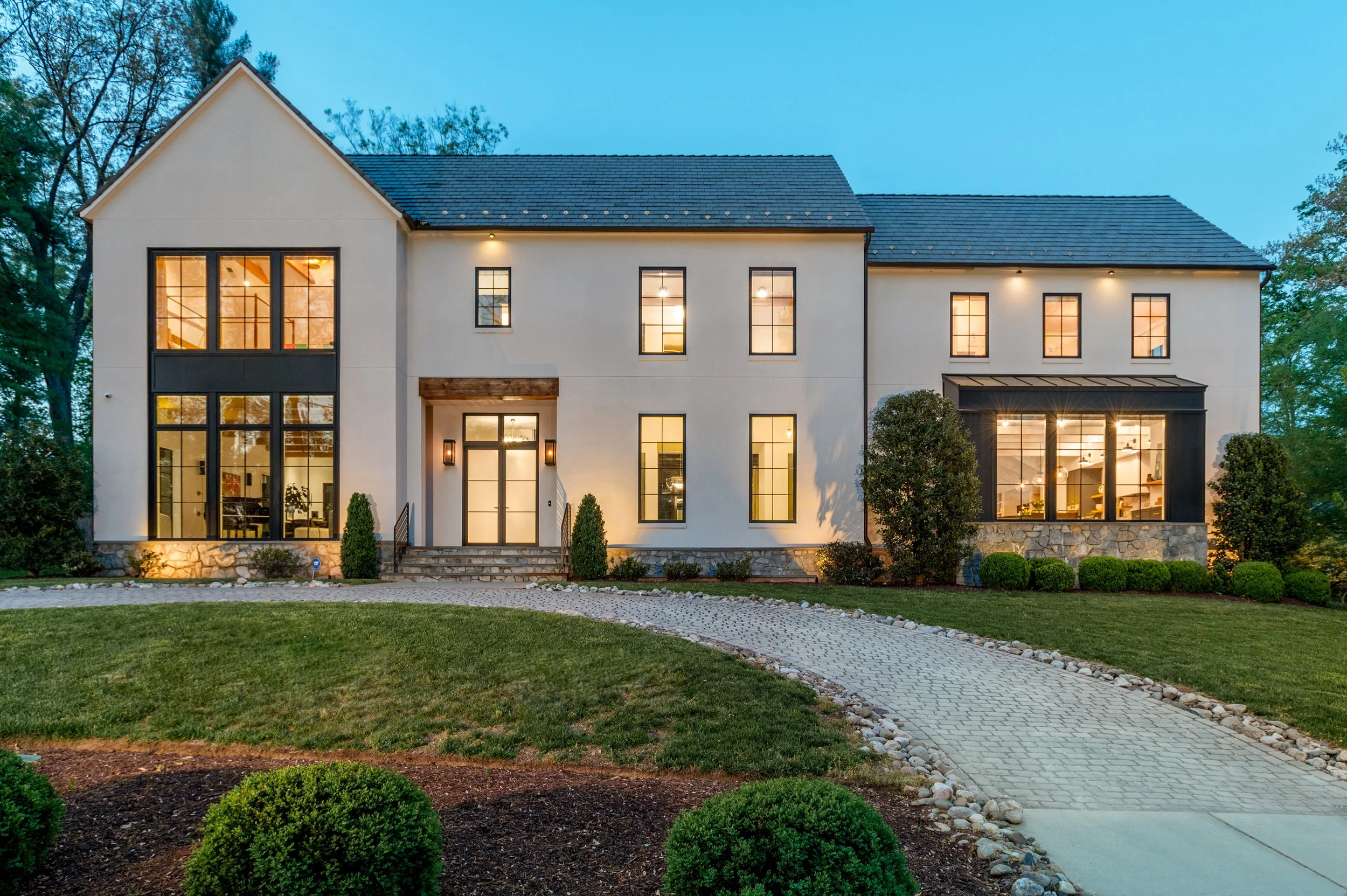 Modern two-story house at dusk with lit windows, front door, and landscaped yard with trees and grass.