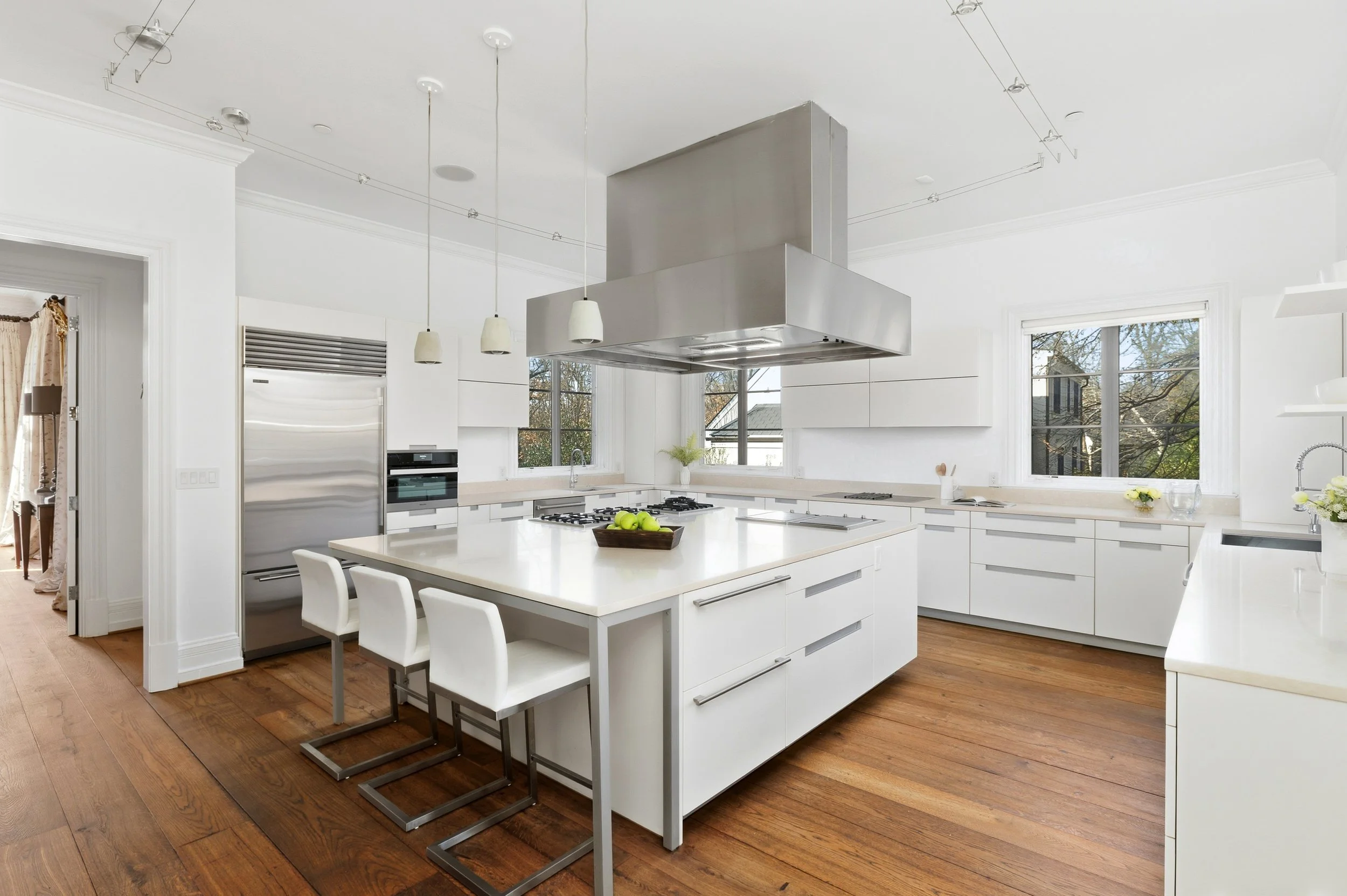 Modern white kitchen with wooden flooring, large island with a bowl of green apples, white chairs, stainless steel range hood, and large windows showing trees outside.