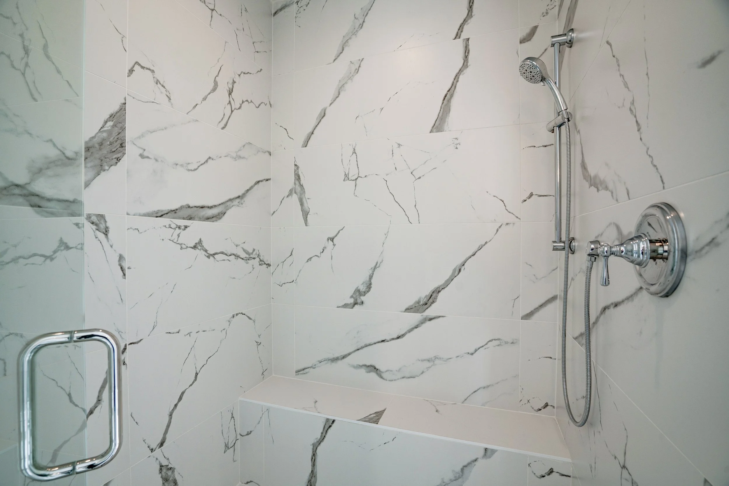 Shower area with large white marble tiles with gray veins, a handheld shower head on a vertical bar, and a chrome control handle.