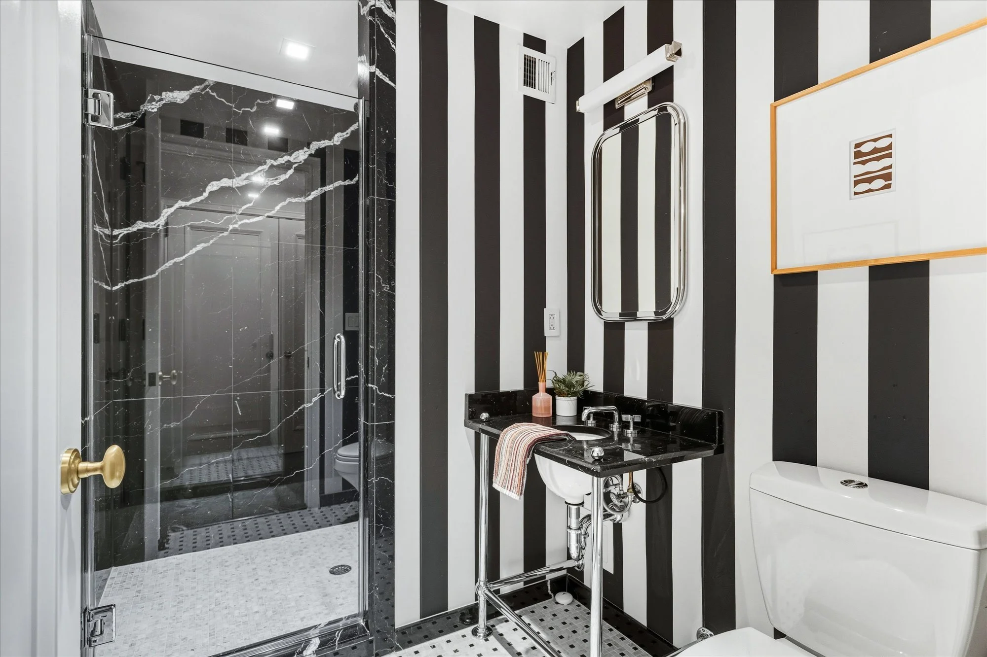 Bathroom with black and white vertical striped wallpaper, a walk-in shower with black marble walls and glass door, a sink with a black countertop and a striped towel, a white toilet, and framed wall art.