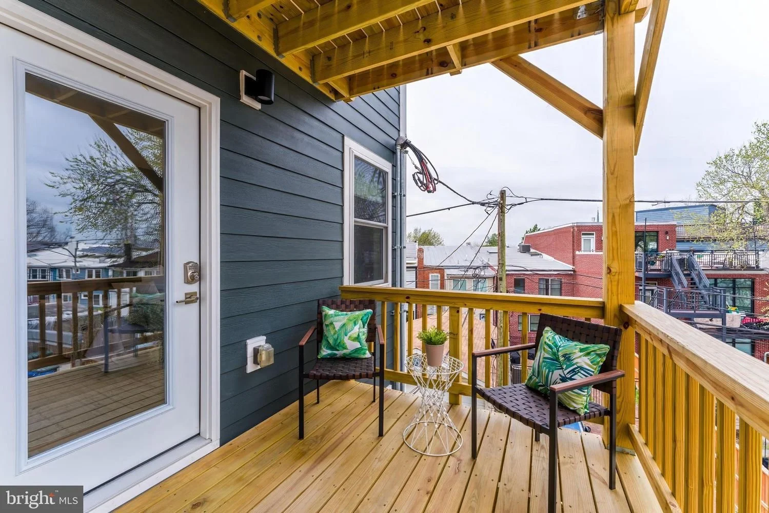 The image shows a small balcony with wooden flooring and railing, attached to a dark blue house. There are two black chairs with green patterned cushions, a small round white table with a potted plant, and a door leading inside. Surrounding buildings