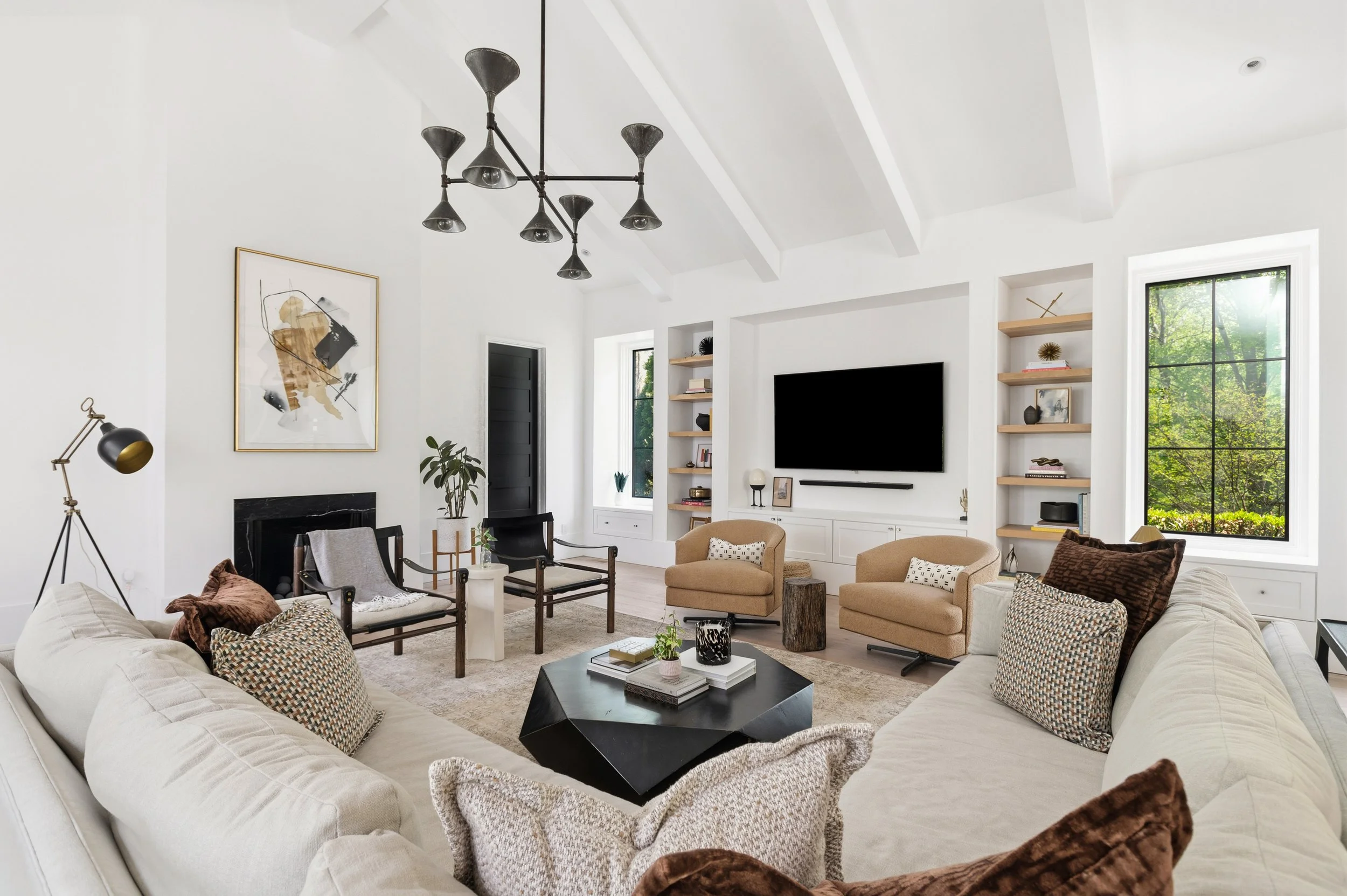 A modern living room with white walls, high ceiling with exposed beams, multiple windows with green trees outside, black and wood shelves, a wall-mounted TV, several beige and patterned armchairs, a black coffee table, and a black chandelier with mul