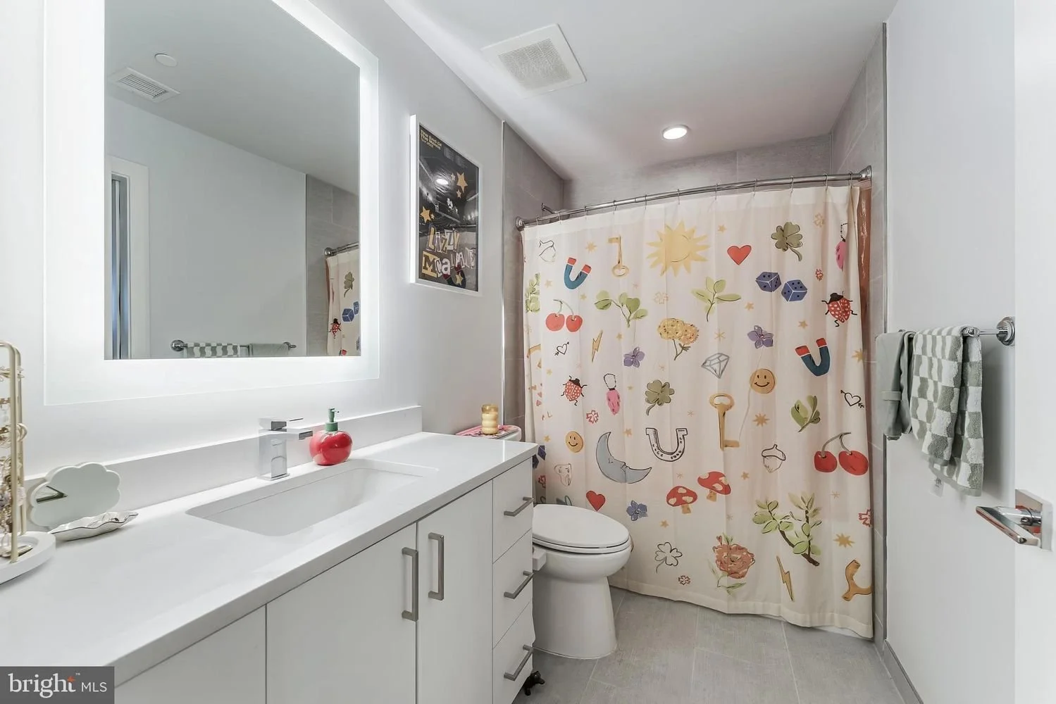A modern bathroom with a white vanity, large mirror, and a colorful shower curtain with playful designs including fruits, animals, and symbols. There are gray towels on a rack, a framed picture on the wall, and various small items on the vanity count