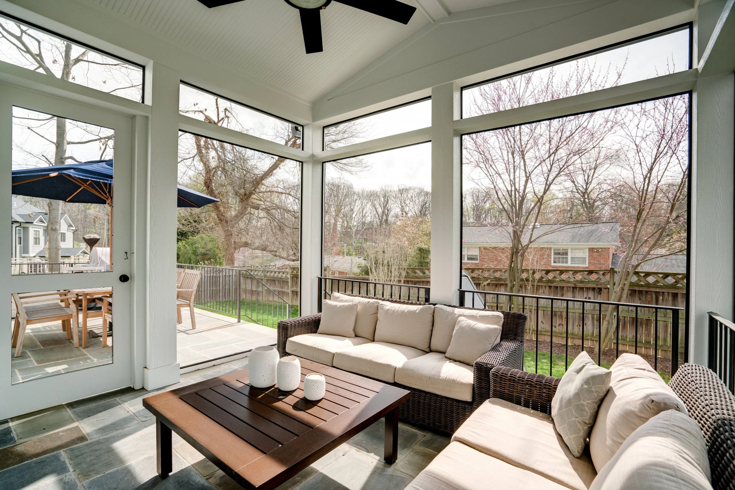 Enclosed porch with outdoor furniture, ceiling fan, and large windows overlooking a backyard with trees and a fence.