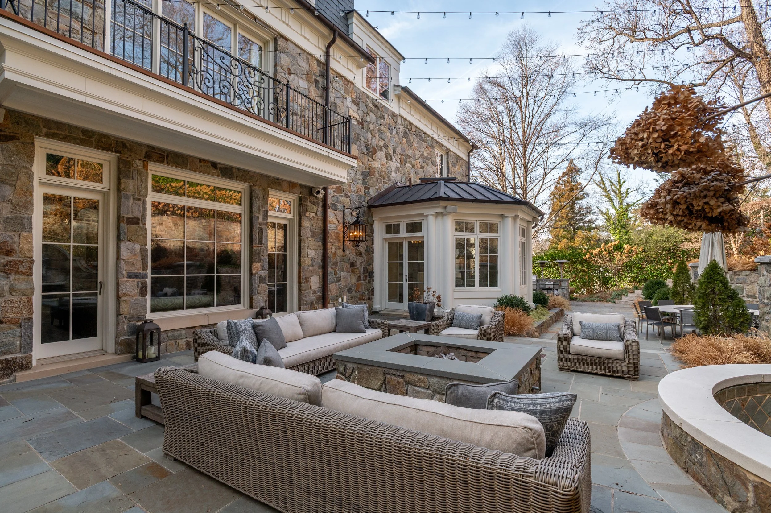 A backyard patio area with outdoor furniture, including beige cushioned sofas and wicker chairs, in front of a stone house with large windows and a bay window. There are string lights hanging above, trees with no leaves, and neighboring houses in the