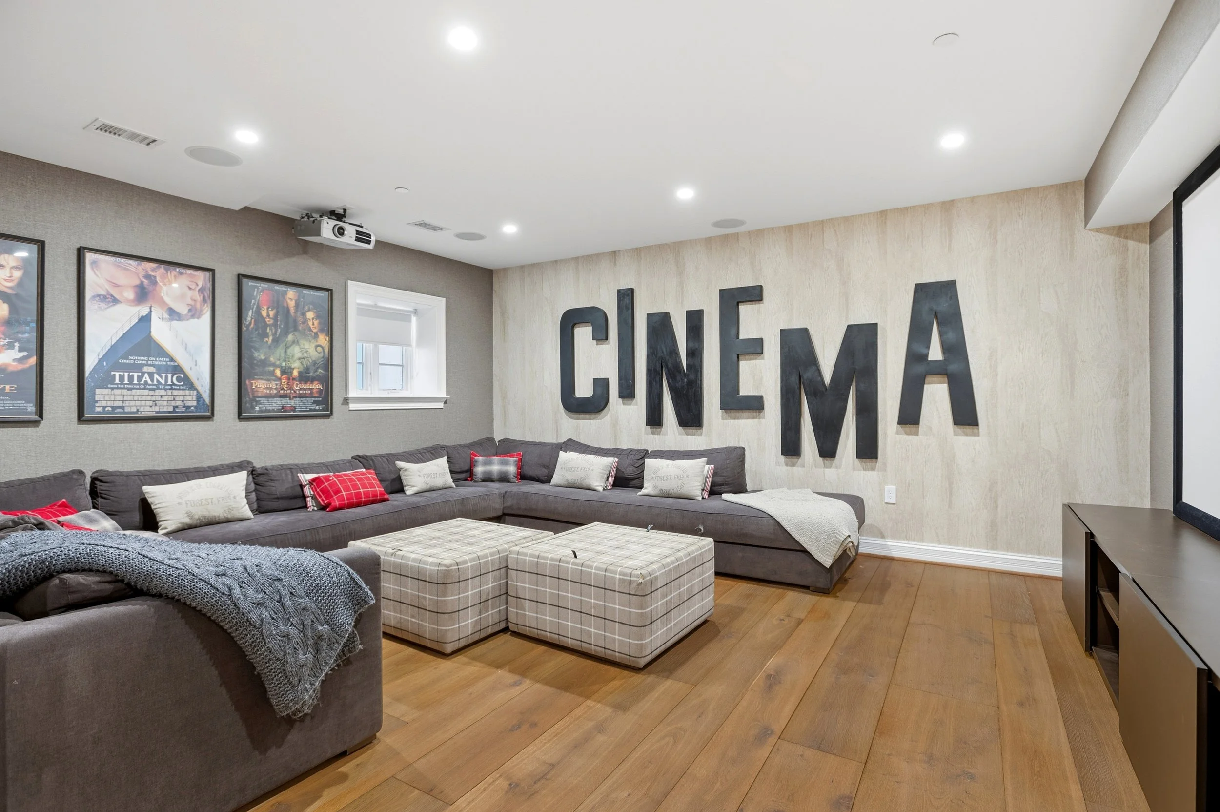 A cozy living room with a large gray sectional sofa decorated with red and white pillows, a checkered ottoman, and a blanket. The wall features movie posters and a large black 'CINEMA' sign, with a window providing natural light.