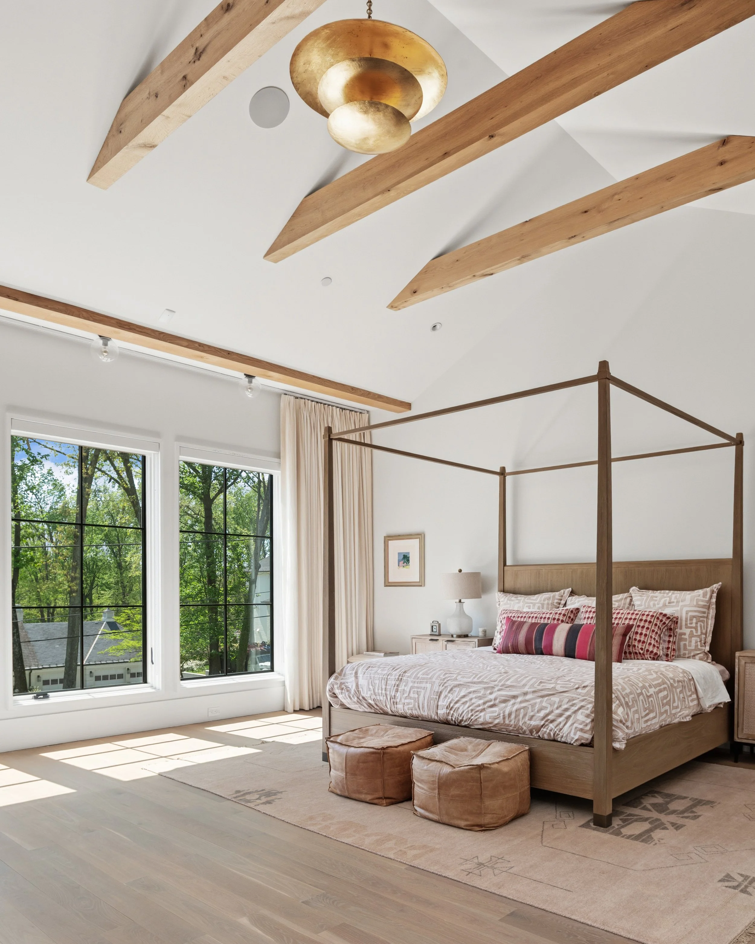A bedroom with a large bed with a wooden frame, multiple patterned pillows, and a beige comforter, adjacent to large windows with white curtains, a white bedside table with a lamp, and an outdoor view of trees and a house, with a chesterfield-style f
