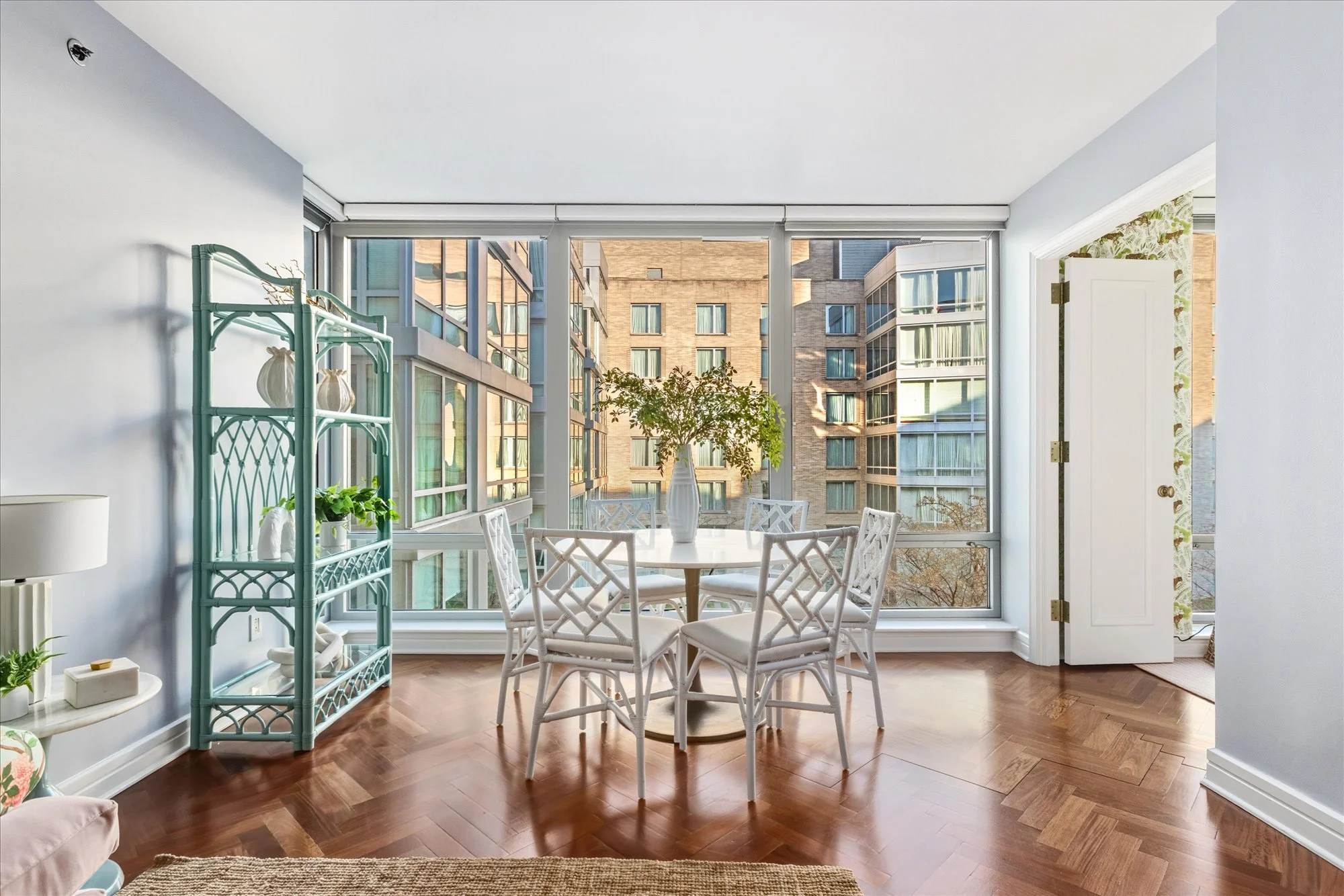 Dining area with white table and chairs, large windows showing neighboring apartment buildings, potted plants, and a light blue shelving unit.