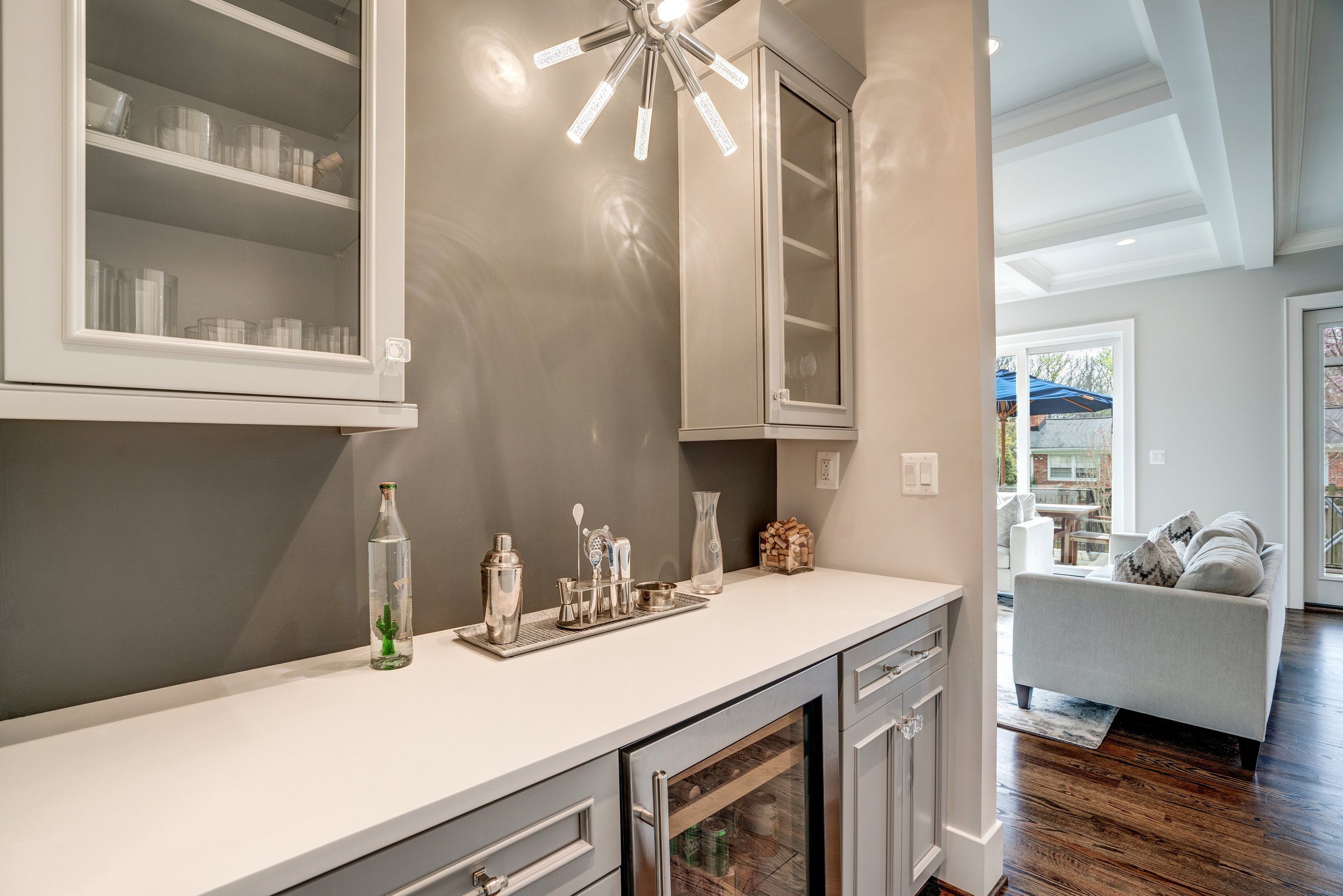 Modern kitchen with white countertops, glass-front cabinets, and a bar area, with a living space and large windows visible in the background.