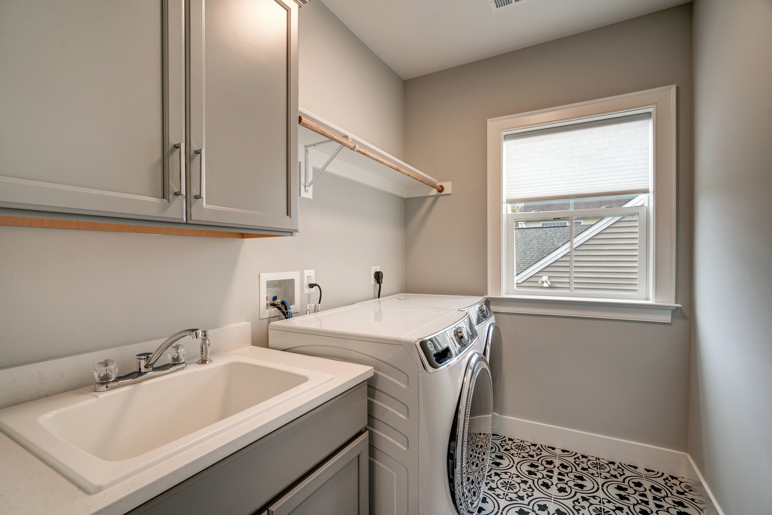 A laundry room with a double basin sink, gray cabinets, a window with a roller shade, and a white washer and dryer side by side. The floor has black and white patterned tiles.