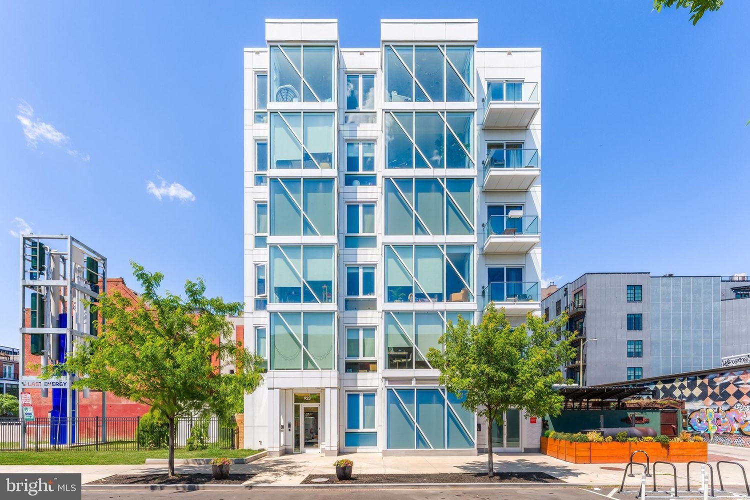 Modern multi-story urban apartment building with large glass windows, small balconies, and two trees in front on a sunny day.