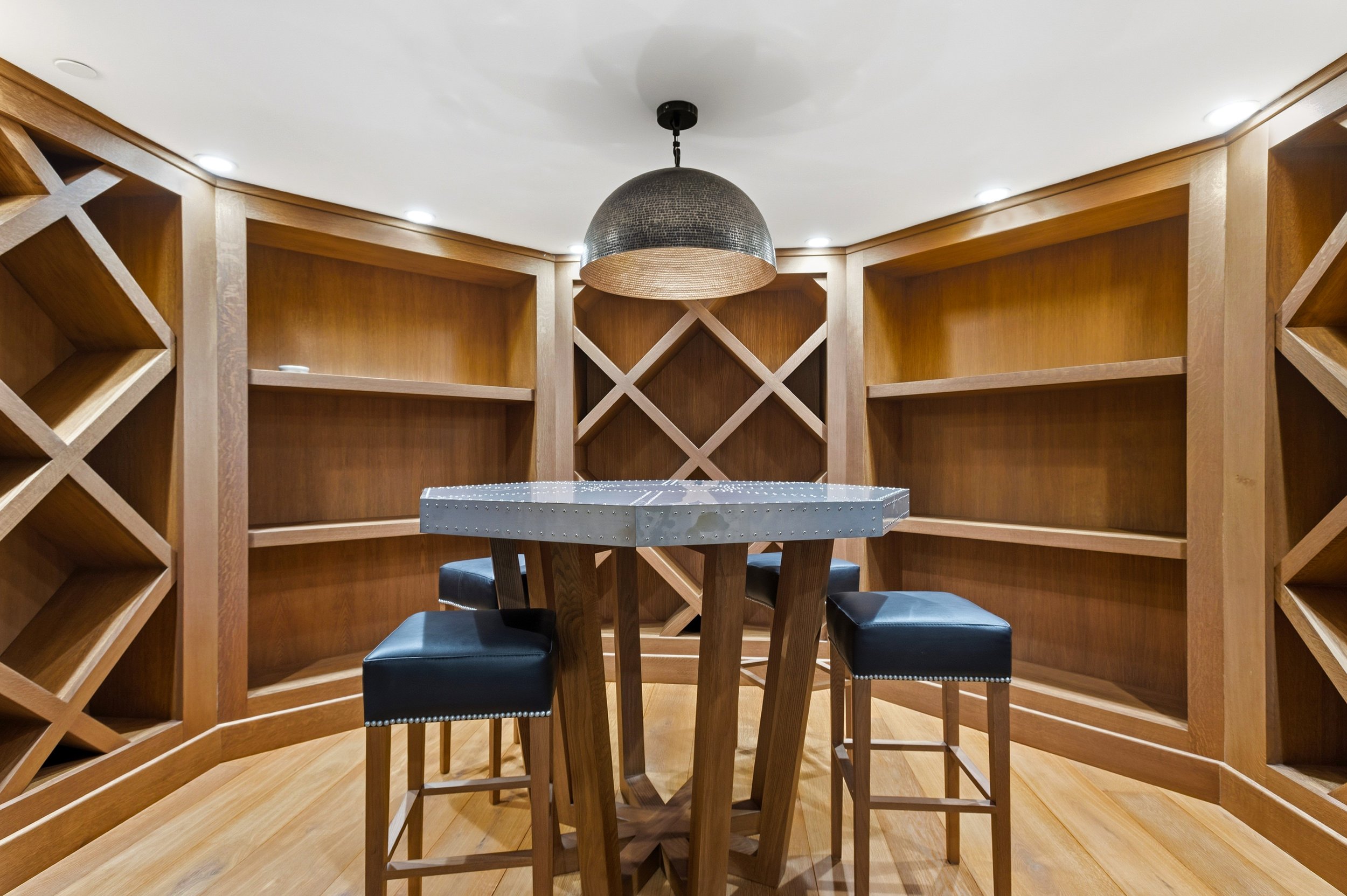 A small wooden dining table with four black leather chairs in a wooden corner wine cellar with wood shelves.
