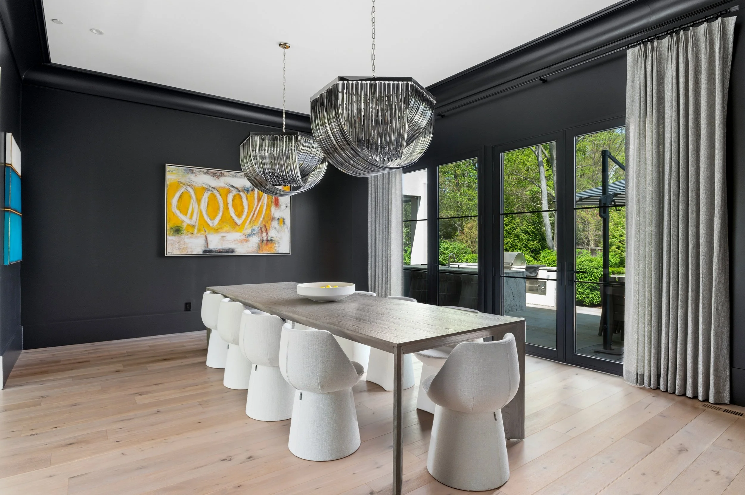 Modern dining room with white chairs, light wood table, and two large glass chandeliers, black walls, abstract painting, and large windows with curtains overlooking greenery.