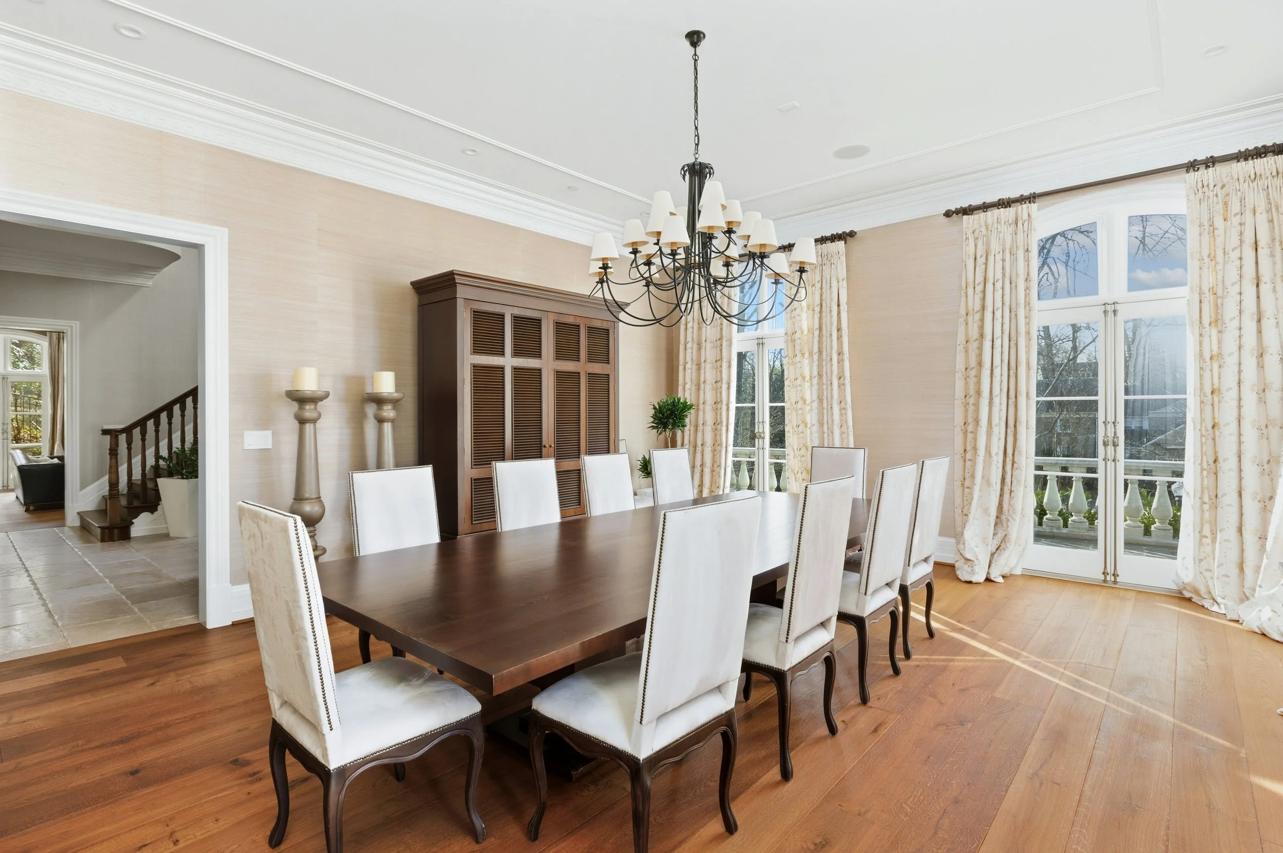 Large dining room with a dark wooden table and white upholstered chairs, a black chandelier with white lampshades, tall windows with floral curtains, a wooden cabinet, and candles on stands.