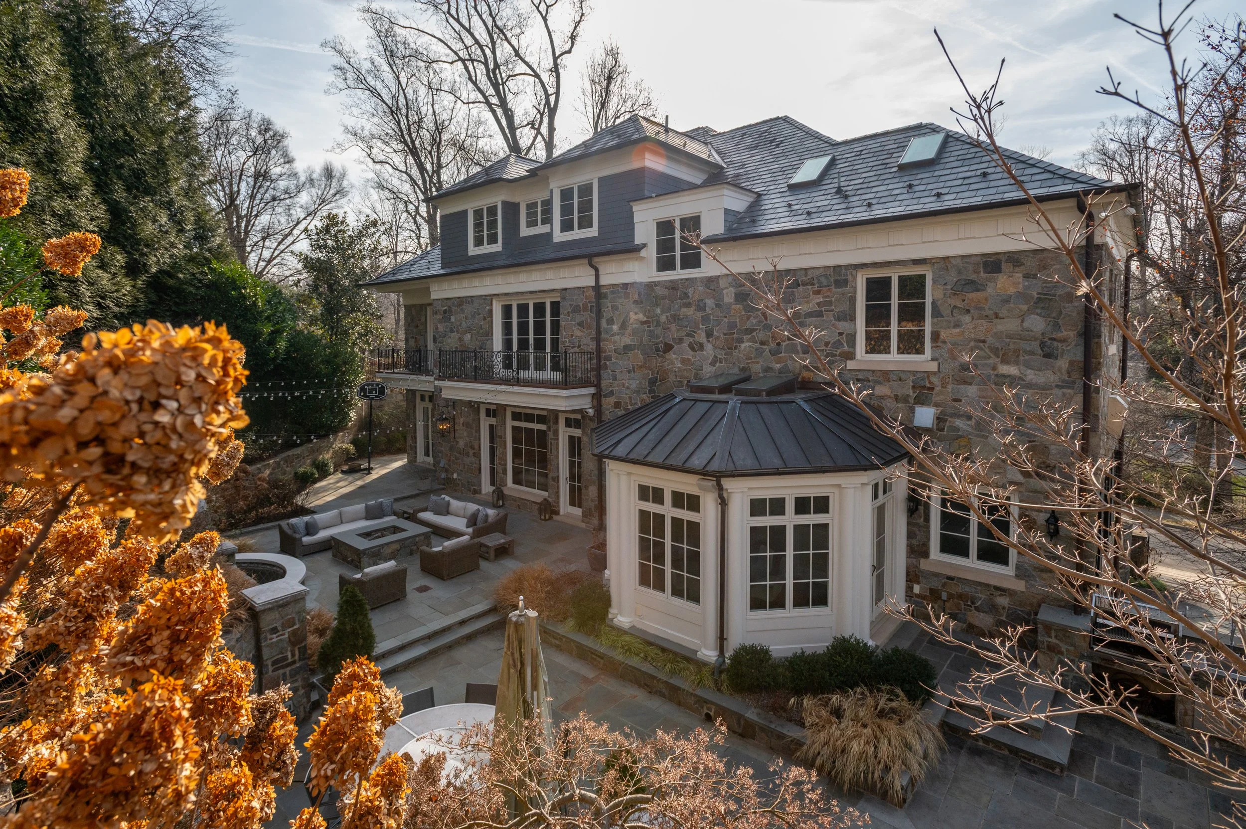 A large multi-story house with stone and blue siding exterior, multiple windows, and a slate roof, surrounded by trees and a landscaped backyard with patio furniture.