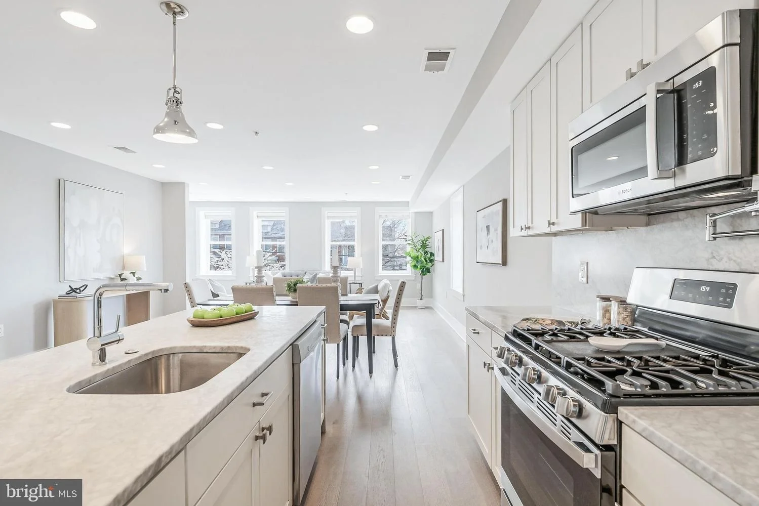 Bright, modern kitchen with white cabinets, stainless steel appliances, marble countertops, and a kitchen island. In the background, a dining and living area with large windows, beige chairs, and a potted plant.