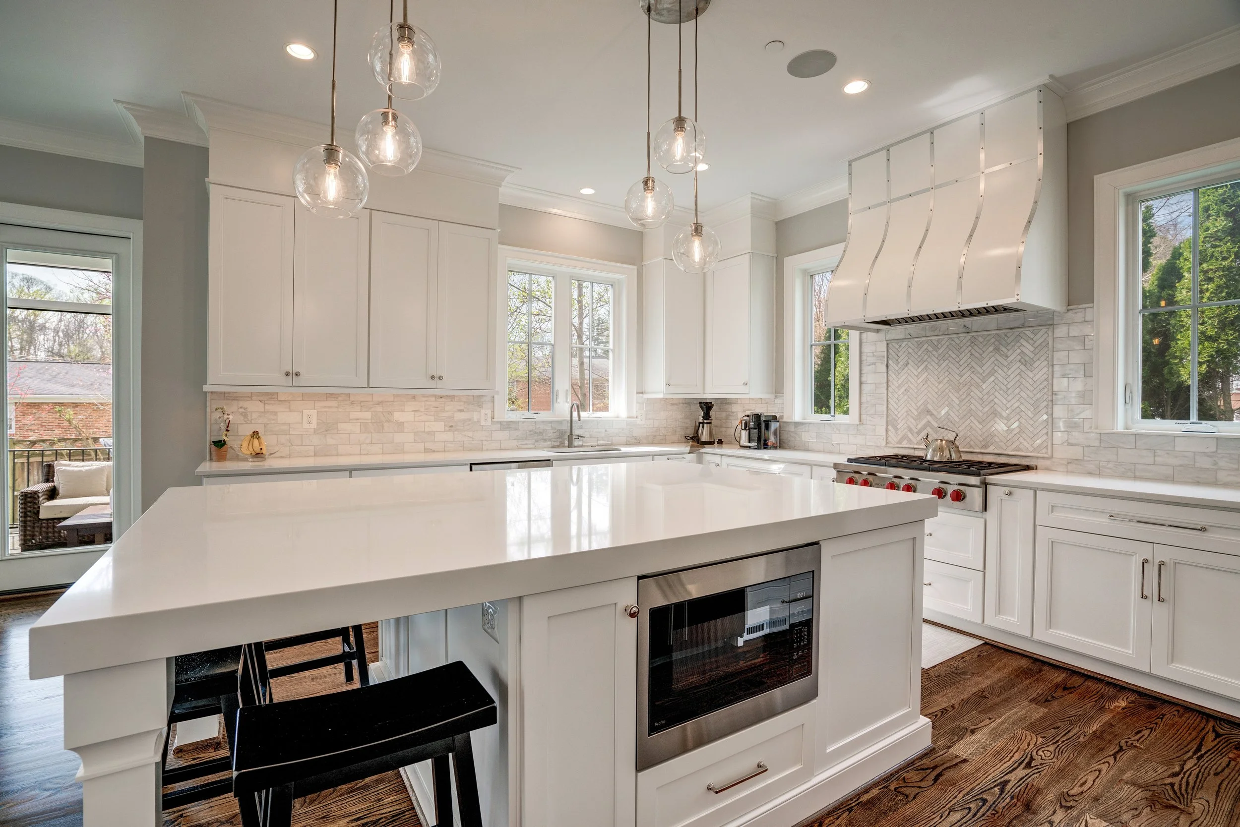 Modern kitchen with white cabinets, a large island with a white countertop, stainless steel appliances, and a decorative backsplash. Pendant lights hang from the ceiling, and large windows let in natural light. There is a view of a patio with outdoor