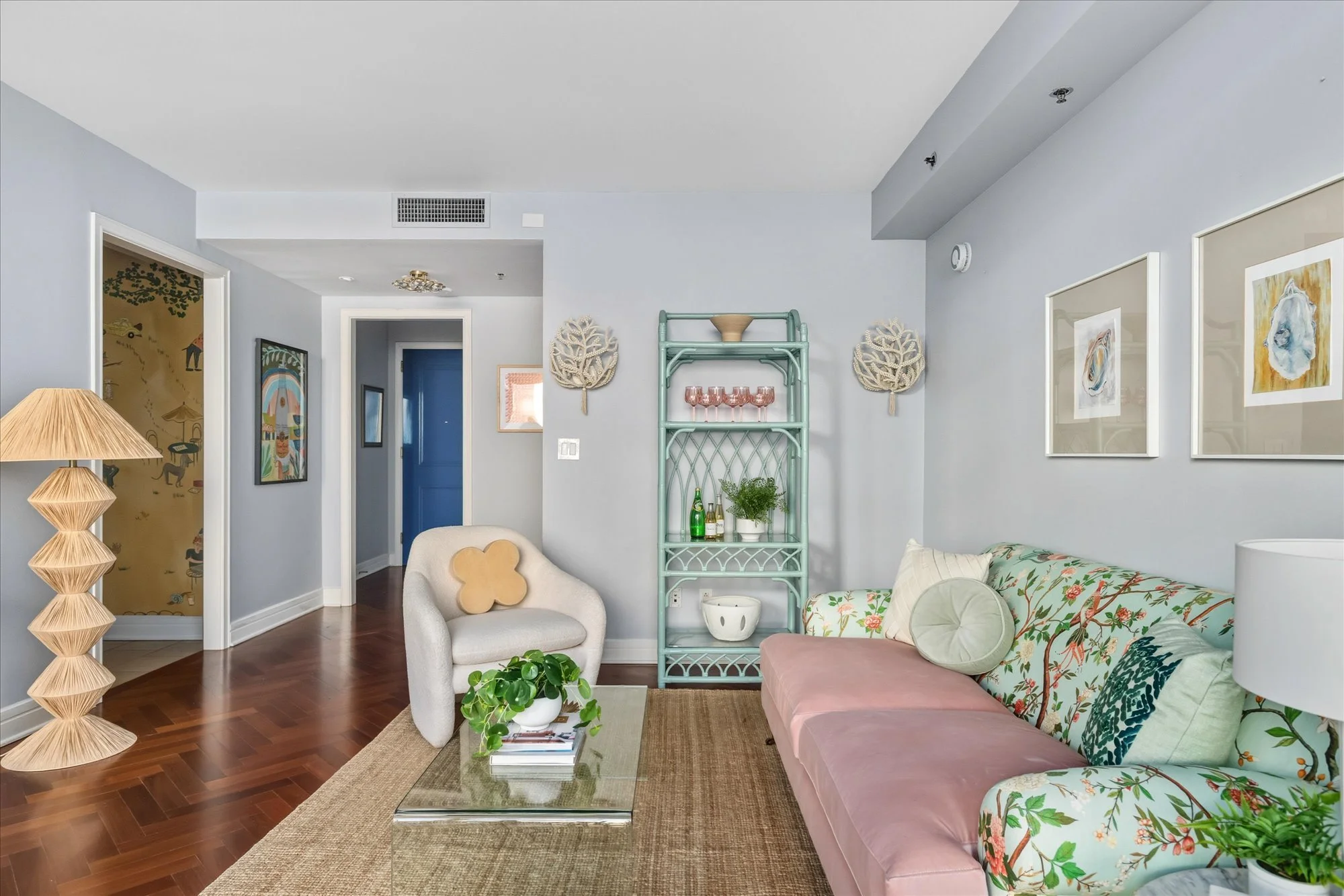Living room with a floral sofa, an armchair with a flower-shaped pillow, a glass coffee table with books and a plant, artwork on the walls, a green shelving unit with wine glasses, and a woven rug.