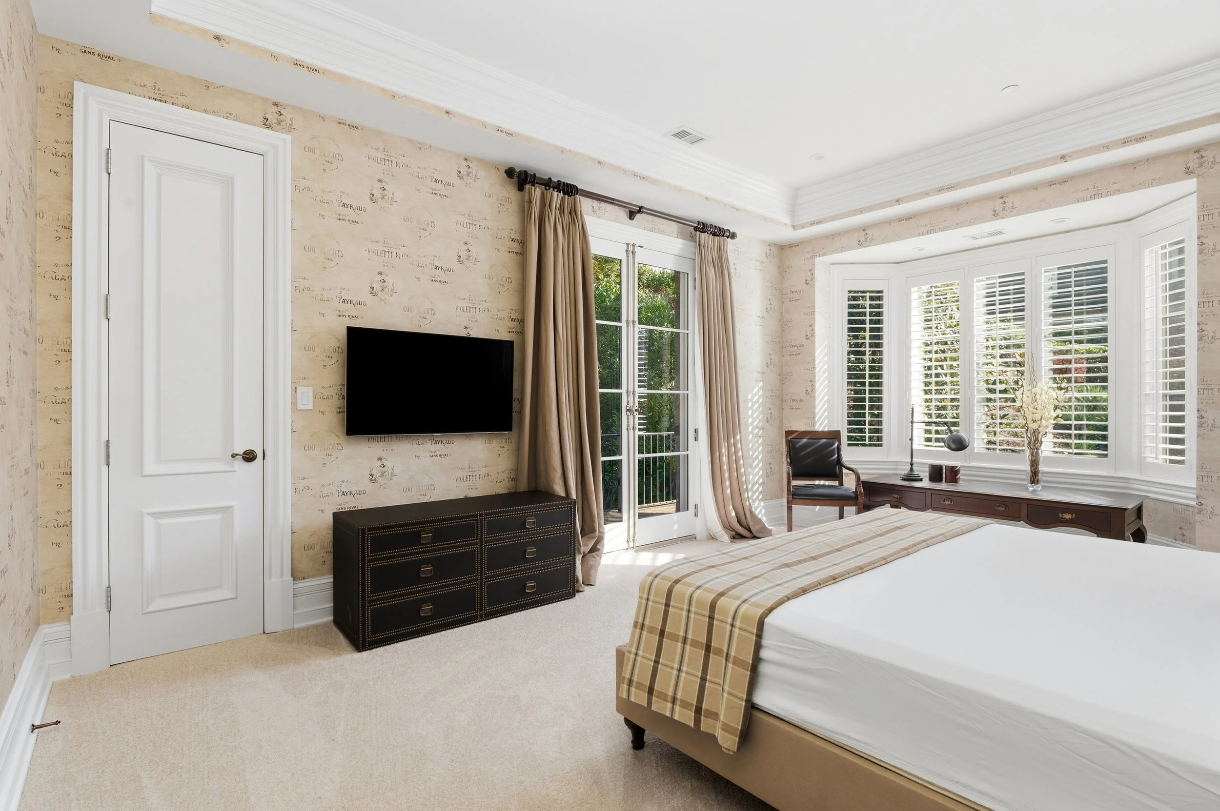 Bright bedroom with large bay window with white shutters, beige walls with a pattern, a white door, a black dresser, and a wall-mounted TV. Bed with a plaid bedspread is visible in the foreground.