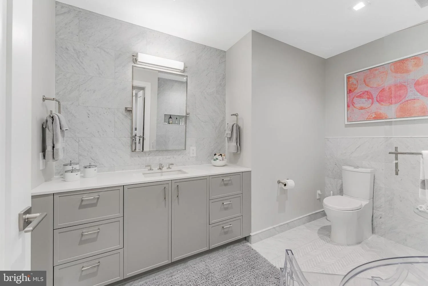 Modern bathroom with light gray cabinets, marble walls, a white toilet, a large mirror, and colorful abstract artwork.