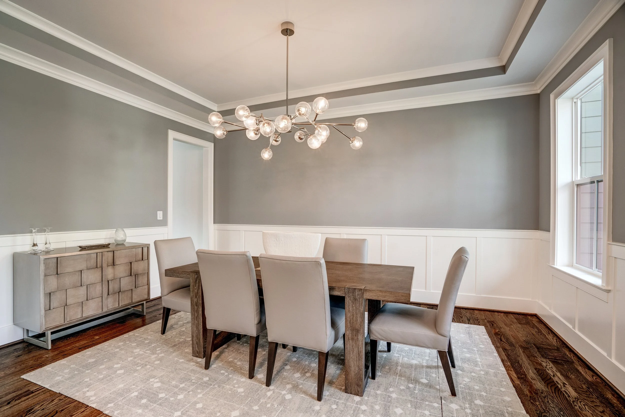 Modern dining room with a wooden table, six beige upholstered chairs, a geometric chandelier, gray walls, white trim, two windows, a wooden sideboard, and a light-colored area rug.